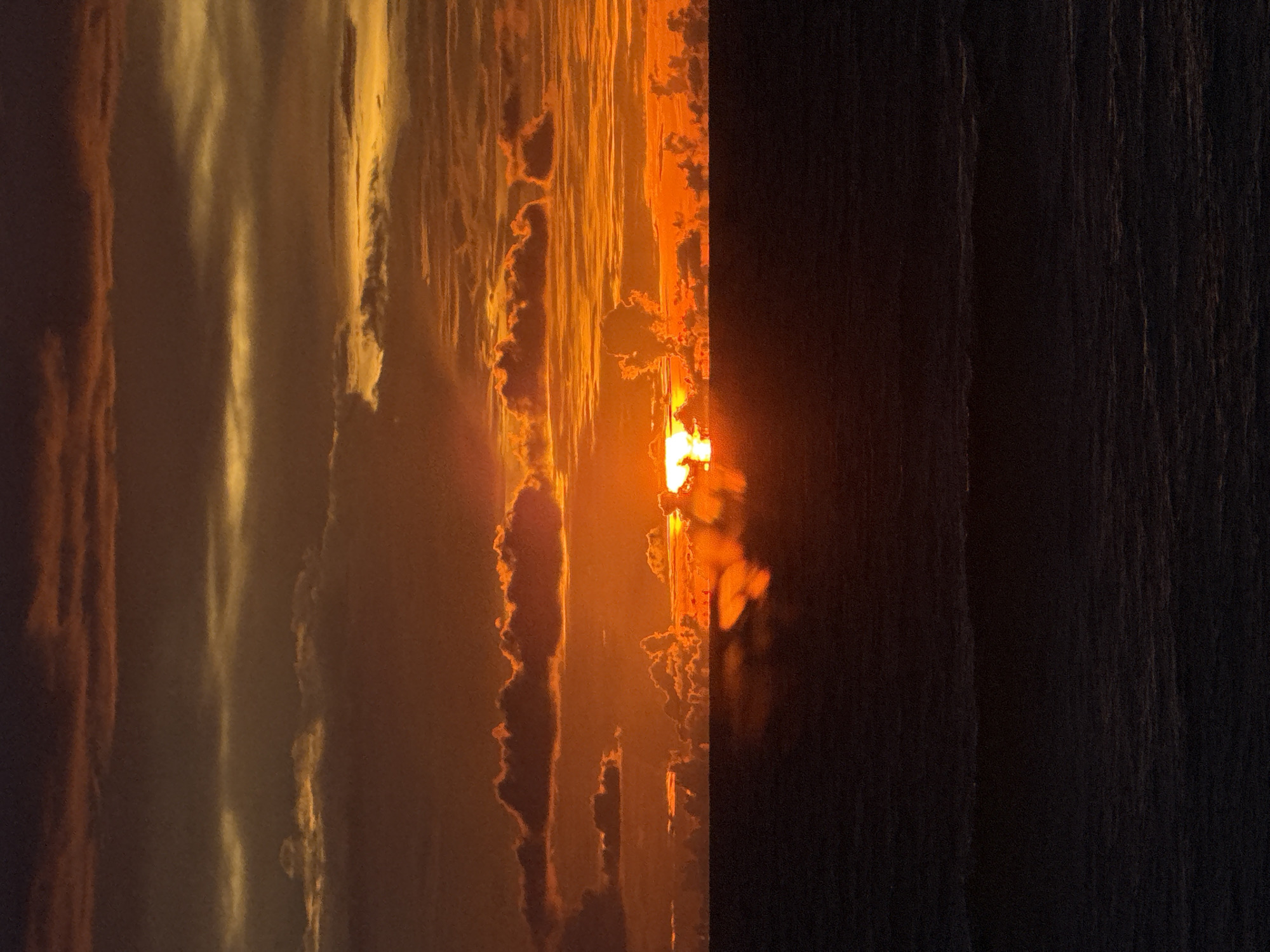 This is a vertical, high-angle, close-up shot of a sunset over a body of water. The sun, a bright orange ball, is setting in the center-right of the frame, casting a warm glow across the sky. Wispy clouds, illuminated by the setting sun, are scattered across the sky, creating a textured and dynamic scene. The water in the foreground is dark and appears to be moving, with visible ripples.
The overall mood of the image is serene and peaceful, evoking a sense of awe and wonder at the beauty of nature. The colors are rich and vibrant, with a strong contrast between the bright orange of the sun and the dark blues and grays of the water. The close-up perspective allows the viewer to appreciate the intricate details of the clouds and the texture of the water.
The time of day is clearly sunset. The weather appears to be clear, with a few scattered clouds. There are no visible people or objects in the frame, making the focus entirely on the natural elements. The location is not specified, but the presence of a large body of water suggests a coastal or lakeside setting. The image is abstract and impressionistic in its style, focusing on the interplay of light and color.