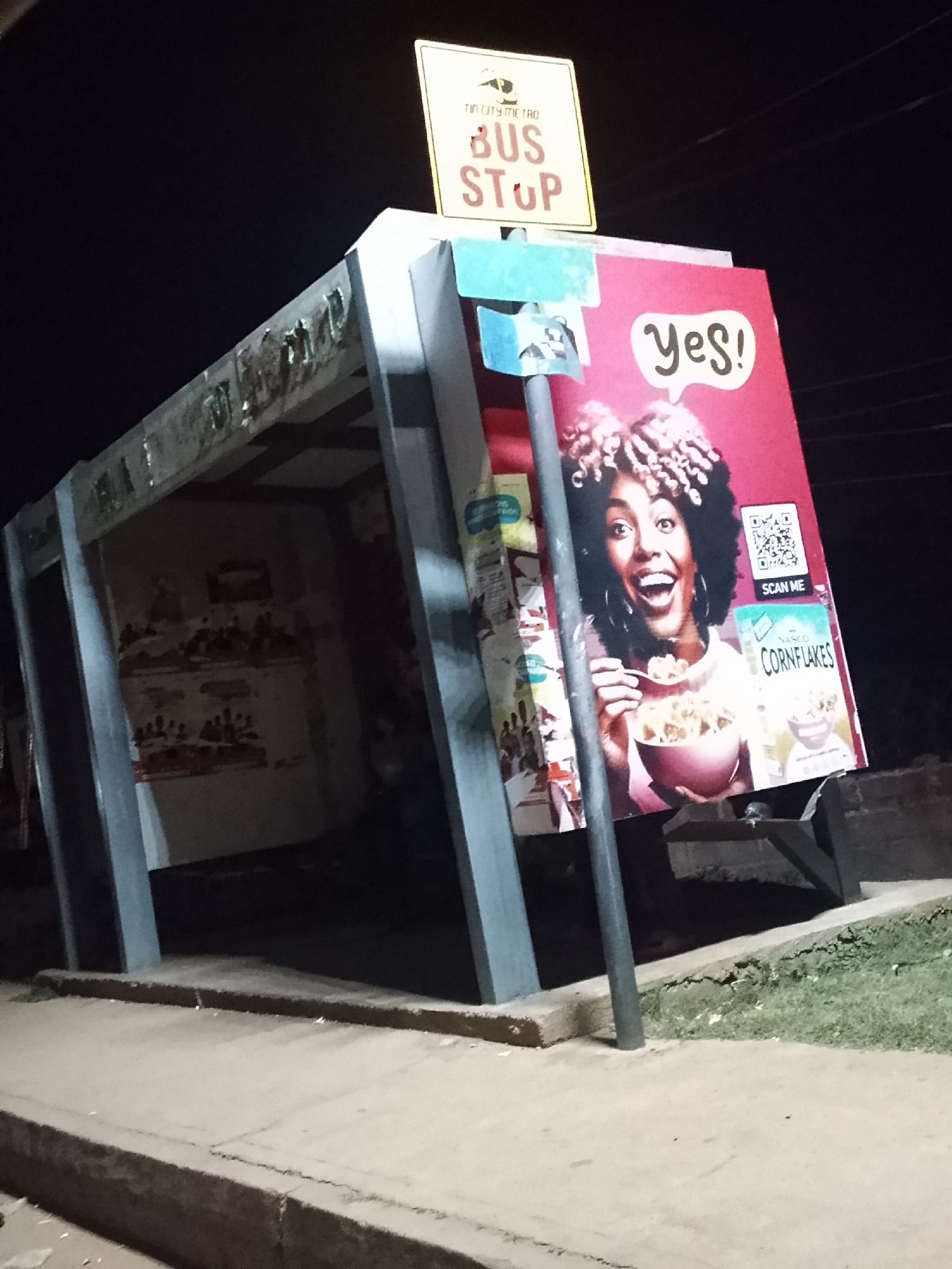 The image shows a bus stop at night. The sign reads "Tin City Metro Bus Stop." The bus stop has a large advertisement for Nasco Cornflakes featuring a woman eating cereal. The location is Bukuru, Nigeria. The bus stop is made of gray concrete and has a roof. The surrounding area is dark and appears to be a sidewalk with grass. There is no one present.