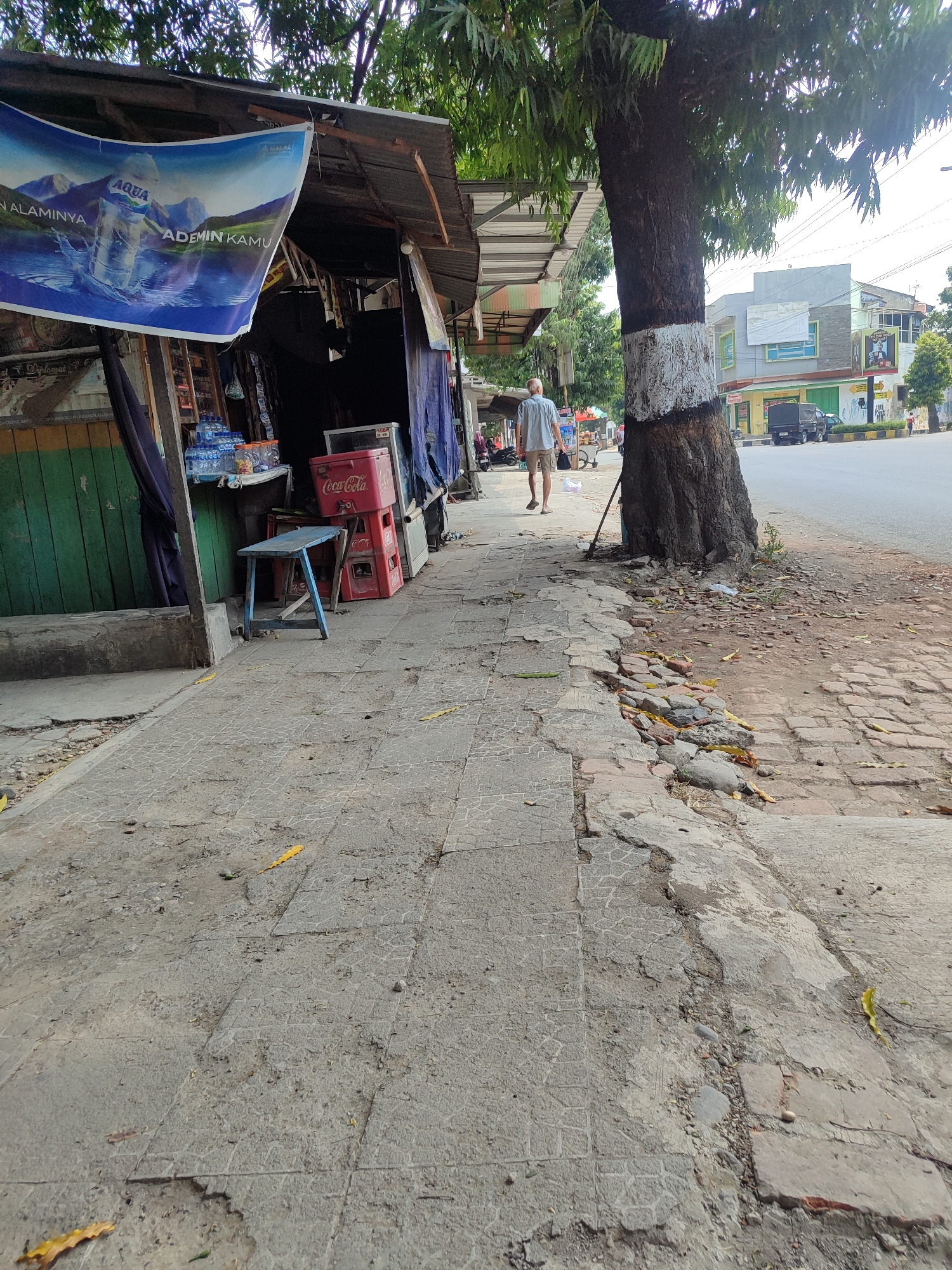 The image depicts a sidewalk in Kagok, Indonesia, adjacent to a small roadside stall. A man, dressed in a light blue short-sleeved shirt and beige shorts, walks away from the camera down the sidewalk. He is wearing flip-flops. The stall has a corrugated metal roof and displays a banner advertising "AQUA" bottled water. A blue stool and a red Coca-Cola cooler are visible in front of the stall, along with stacks of bottled water. The sidewalk is made of concrete tiles and shows signs of wear and damage, with some areas of cracked and uneven pavement. A large tree with a trunk painted with white markings stands near the sidewalk, its branches extending over the scene. In the background, there is a road with a few vehicles and buildings lining the street. The overall environment suggests a warm, sunny day.