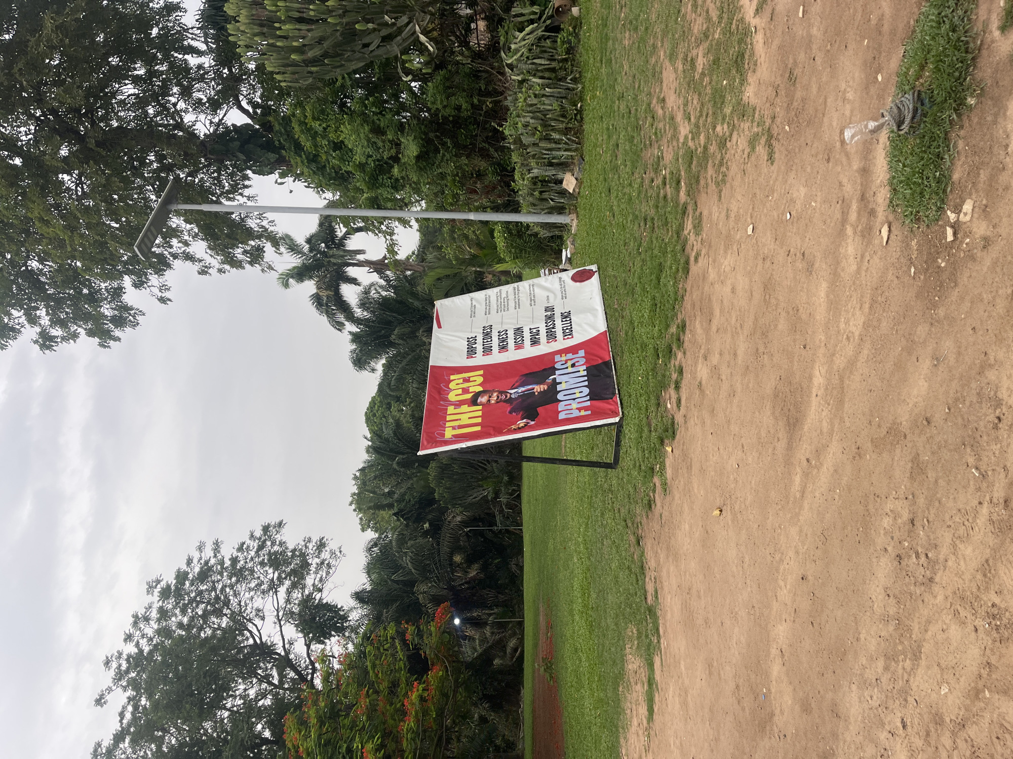 The image shows a poster standing on a green lawn next to a dirt road in Galadimawa Village, Nigeria. The poster advertises "THE CCI PROMISE" and includes a picture of a man in a suit. The background consists of trees and a cloudy sky. A modern street light is visible in the mid-ground. A discarded plastic bottle wrapped in rope is sitting on the edge of the lawn, where it meets the dirt road.