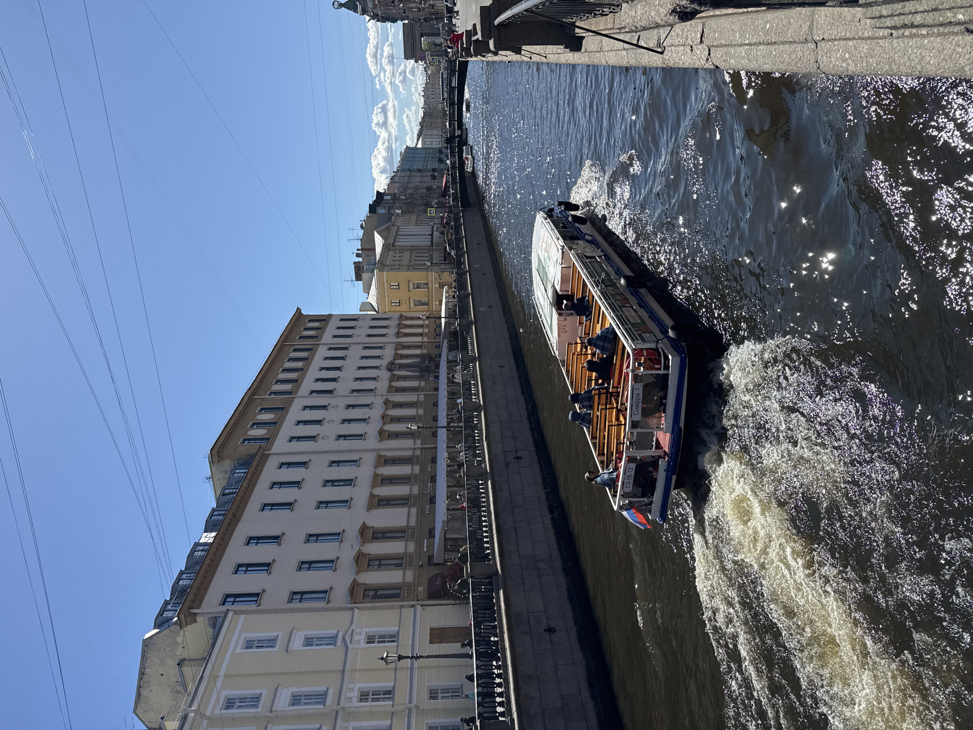 The image depicts a boat navigating a canal in Saint Petersburg, Russia, as evidenced by the architectural style of the buildings lining the canal. The boat, occupied by several passengers, is moving through the water, creating a wake. The canal is bordered by a stone embankment. The sky is clear and blue, with electrical lines visible overhead. The buildings adjacent to the canal display a classic, multi-story design with numerous windows. Some pedestrians are visible on the embankment. The scene is bathed in bright sunlight, causing reflections on the water's surface.