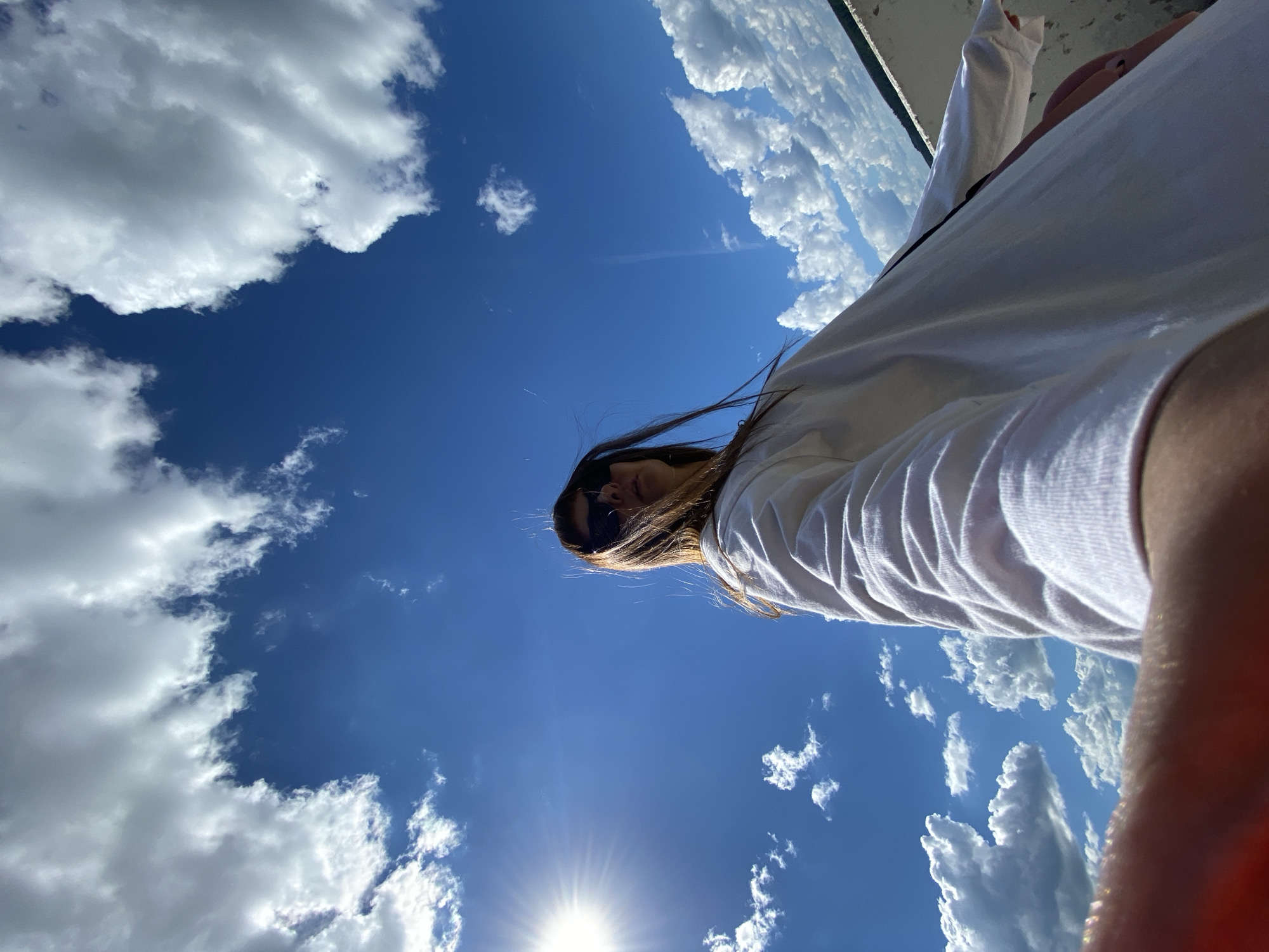 A low-angle, wide shot captures a young woman facing away from the camera, her face partially obscured by sunglasses and her long brown hair blowing in the wind. She is wearing a long-sleeved white shirt and appears to be holding out her arm to take a selfie. The background is a bright blue sky filled with scattered white clouds, and the sun is shining brightly on the left side of the frame. A white railing or wall is visible along the bottom right edge of the image. The scene suggests an outdoor setting, possibly on a rooftop or elevated area, with a sense of open space.
