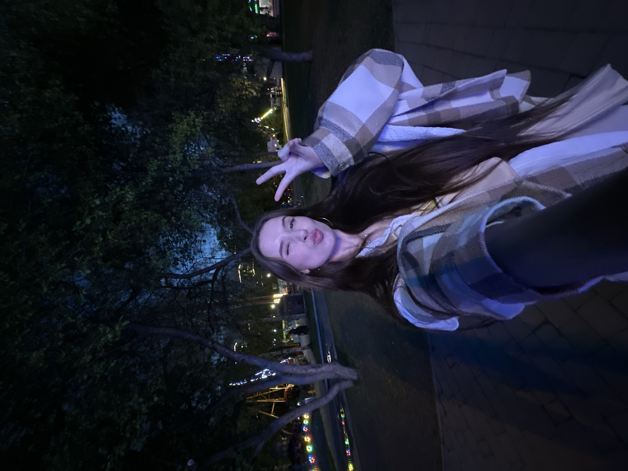 A young woman takes a selfie at night in Yekaterinburg, Russia. She wears a plaid shirt and makes a peace sign with her fingers, pursing her lips. The background features trees, streetlights, and what appears to be an amusement ride with colorful lights. The scene indicates an urban park or recreational area in the city.