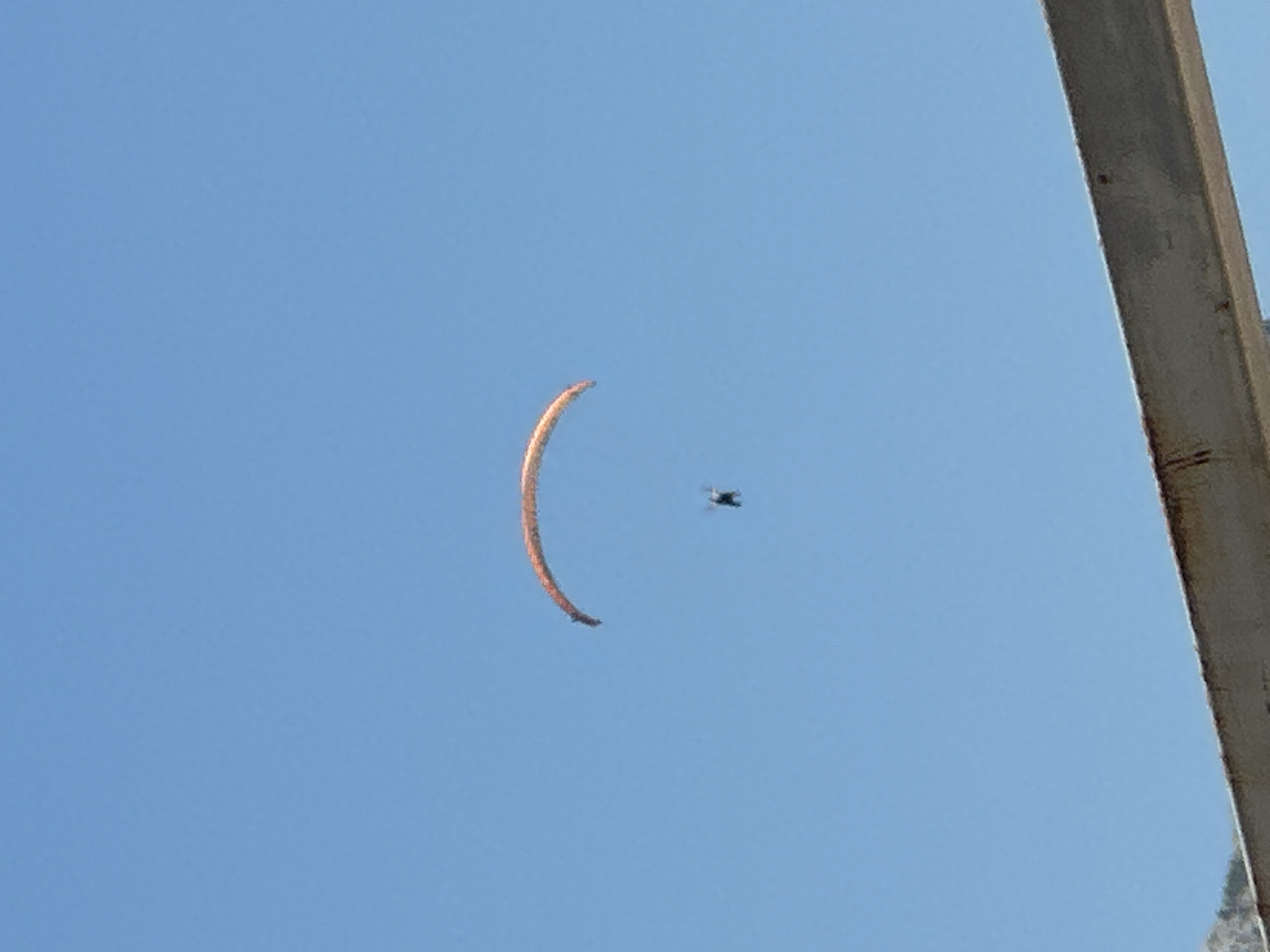 In the city of Boreti, Montenegro, a paraglider is captured against a clear blue sky. The paraglider, an individual suspended beneath a colorful wing, is mid-flight, appearing as a dark silhouette against the vast expanse of the sky. The paraglider's wing is an orange-red arc, gracefully curved and taut, catching the unseen air currents. Below, a portion of a weathered, metallic structure, possibly a bridge or a building's overhang, occupies the bottom of the frame, its rustic texture contrasting with the smooth, uninterrupted sky. The overall scene conveys a sense of freedom and adventure, with the paraglider dwarfed by the immensity of the natural environment.