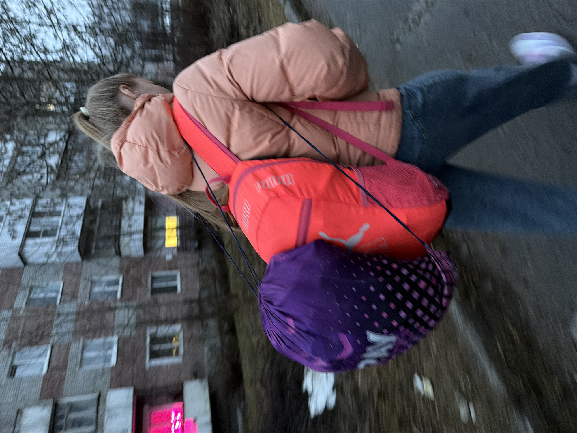 A young person is walking outdoors. They are wearing a peach-colored puffer jacket, blue jeans, and white sneakers. A red Puma backpack is on their back, and a purple and black drawstring bag is attached to it. The background includes a building with multiple windows. The building is located in the city of Berezniki, Russia. The person is in motion.