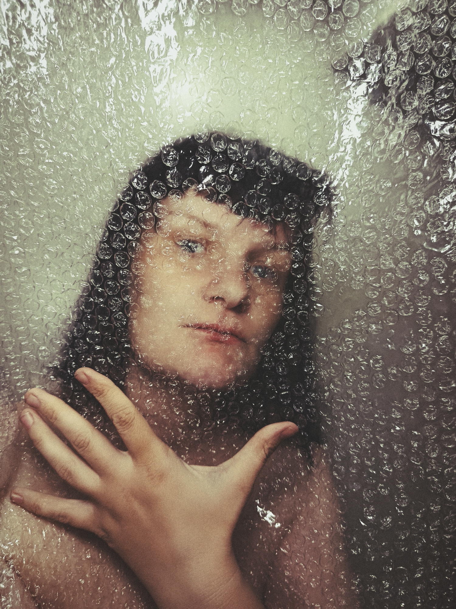 A portrait photograph captures a person, likely female, positioned behind a sheet of bubble wrap that covers the entire frame. The subject has dark, shoulder-length hair with bangs and appears to look directly at the viewer with light-colored eyes. Her facial expression is somewhat neutral, possibly pensive or curious, with slightly parted lips. A hand, with fingers spread, is pressed against the bubble wrap in the lower left foreground, creating a tactile impression against the textured surface.
The bubble wrap acts as a translucent, distorting filter, fragmenting light and obscuring the background, which appears as an indistinct, softly lit area. This material gives the image a unique, grainy texture and visual quality. The overall color palette is muted, dominated by desaturated greens, grays, and warm skin tones, contributing to a contemplative or introspective mood. There is no discernible text within the image. The scene suggests an indoor setting in Фоки, Russia, and the activity implies an intentional pose exploring themes of confinement, protection, or perception through a barrier.