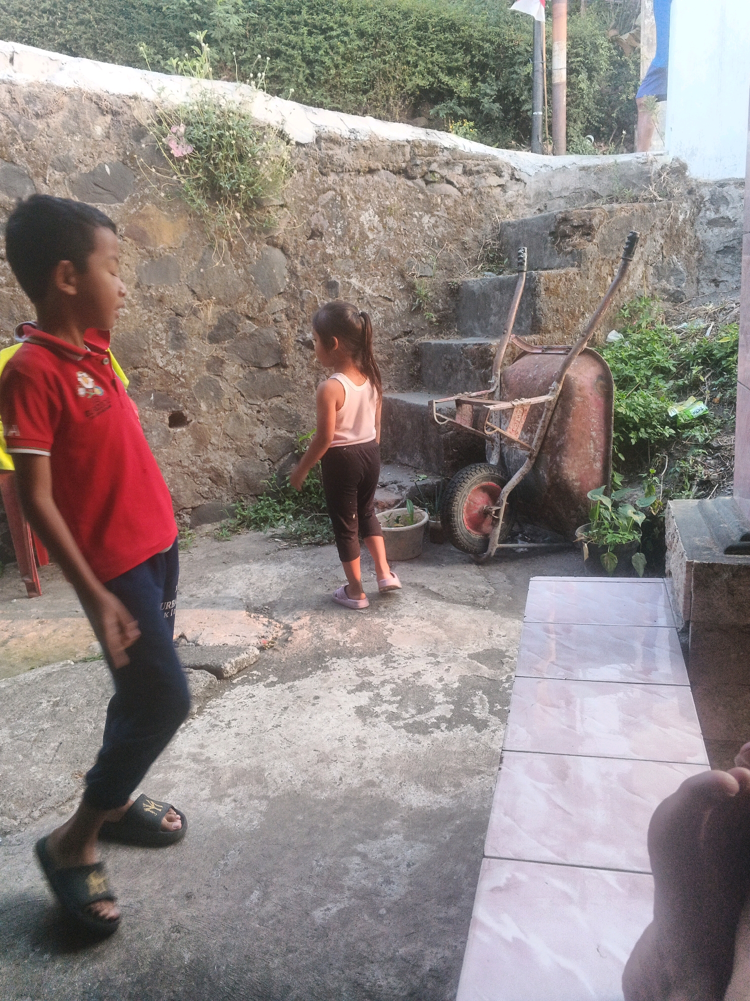 In Keji, Indonesia, two children are present in an outdoor, slightly overgrown area. A young boy, wearing a red polo shirt and dark pants, is walking on a concrete path. To his right, a young girl, dressed in a light pink tank top and dark pants, stands near a rustic wheelbarrow. The wheelbarrow is positioned on a dirt and gravel surface, adjacent to a set of worn, stone steps. A rough stone wall, partially covered in greenery, forms a backdrop. Beyond the wall, dense green foliage suggests a natural setting. A flag on a pole is visible in the upper right quadrant. The overall scene is utilitarian and appears to be an outdoor workspace or yard.