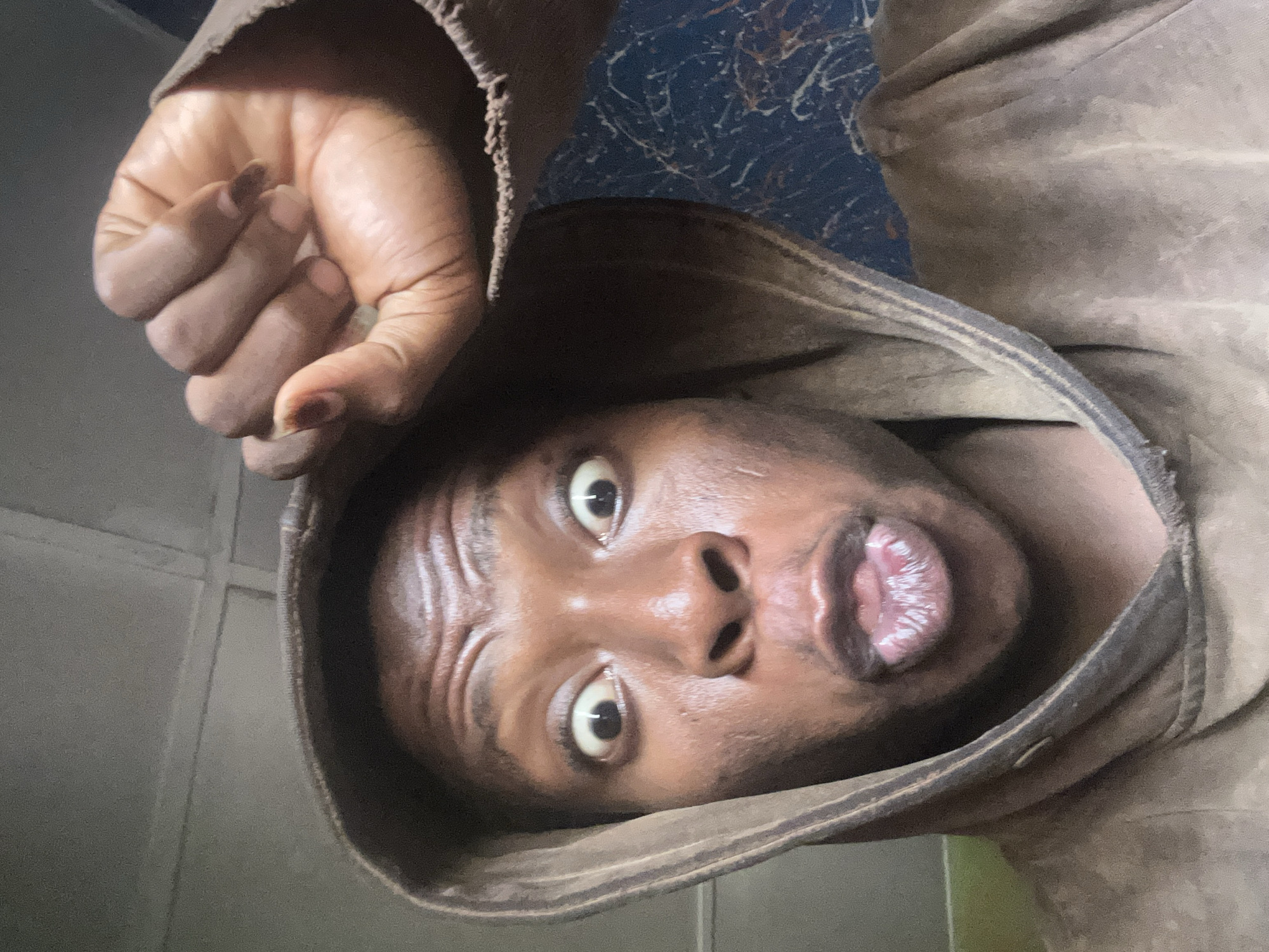 A man with dark skin is centered in the frame, his face angled towards the camera with wide eyes and pursed lips. He wears a brown hooded garment. His right hand, clenched into a fist, is positioned above and to the left of his head. The background is indistinct, but appears to include a dark blue surface with white streaks and a light-colored tiled wall. The location is not known.