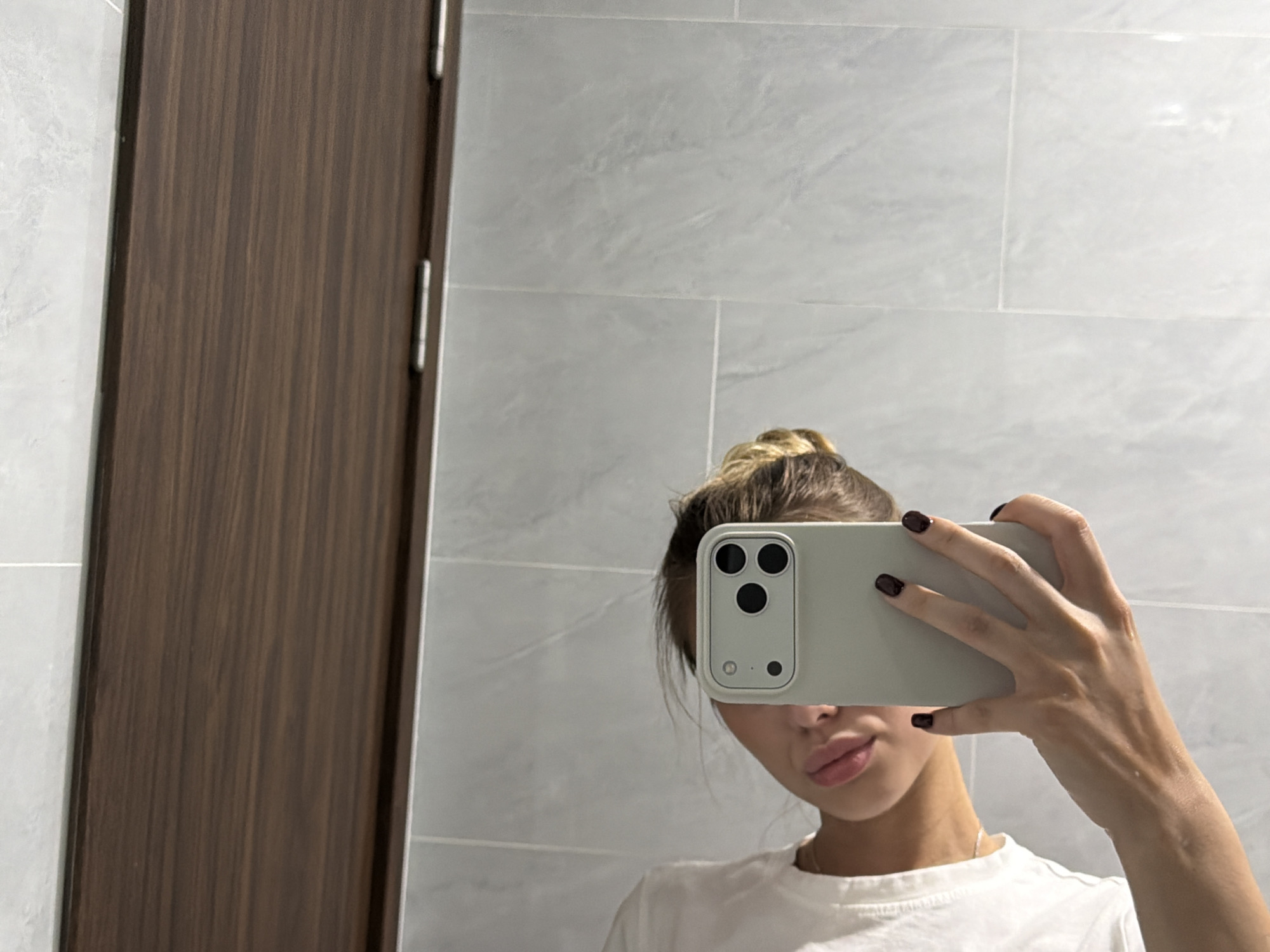 The image is a selfie taken by a young woman in what appears to be a bathroom. She is holding a smartphone to take the photo, which is partially obscuring her face. The background consists of light-colored tiled walls and a wooden door. The woman has her hair tied up in a bun and is wearing a white t-shirt. Her nails are painted a dark color. The lighting suggests it might be daytime, possibly indoors with artificial light. There is no visible text in the image. The overall scene is casual and personal, capturing a moment of self-documentation. The presence of a tiled wall and a wooden door are common elements found in bathrooms in residential or hotel settings. The clean and minimalist background does not provide specific cues about the exact location beyond it being an interior space. The woman's expression, as much as can be seen, appears neutral.