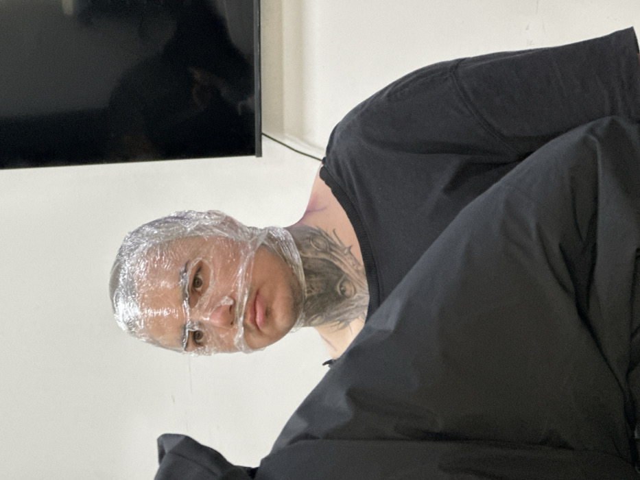 The image presents an indoor scene featuring an individual, likely an adult male, with short hair. His head and face are tightly enveloped in clear plastic cling film, creating indentations over the bridge of his nose, around his mouth, and across his forehead. Visible dark tattoos adorn his neck, prominently featuring an eye motif on the anterior throat. He wears a dark, short-sleeved t-shirt and maintains a neutral expression, gazing slightly towards the viewer's right. In the lower left foreground, a portion of a black garment, consistent with a jacket or coat, partially obstructs the primary subject. The background is a plain white wall. To the right of the primary subject, a dark, rectangular flat-screen monitor or television is mounted on the wall, with a black cable descending from its lower edge. The scene is uniformly illuminated by what appears to be artificial light. The specific location within the city of Rostov-on-Don, Russia, is not discernible from the visual evidence provided in the image.