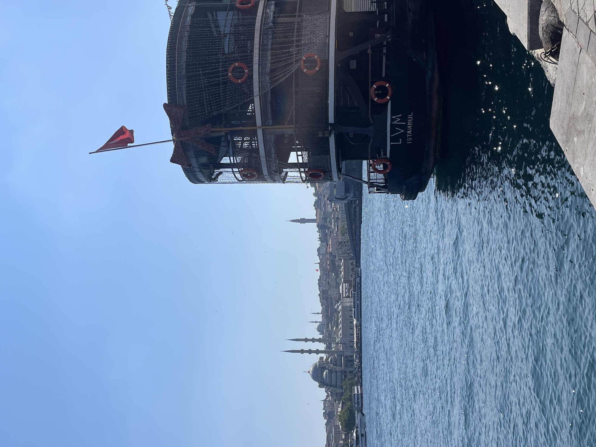 An outdoor, full, eye-level shot captures the front portion of a dark-colored, multi-deck tour boat docked on the right side of the frame. The boat is decorated with strings of small, white lights and features several life preservers. The boat's name, "LVM ISTANBUL," is visible on its stern. In the background, across a body of blue water, is the Istanbul skyline, dominated by a large mosque with multiple minarets and domes. Additional buildings and minarets are scattered across the cityscape, with a bridge visible in the distance. The sky is clear and bright blue. The water is rippled and reflects the sunlight. The scene appears to be a sunny day. The location is likely the Golden Horn or Bosphorus strait in Istanbul, Turkey, with the mosque being the Süleymaniye Mosque.