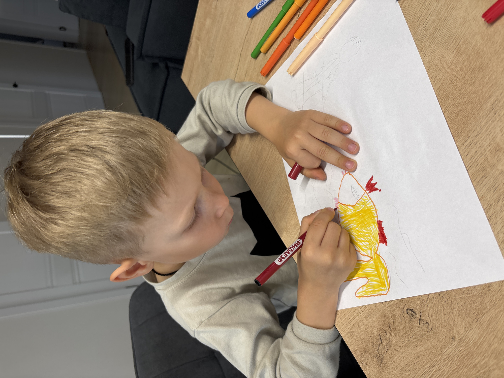 A high-angle shot captures a young child, likely a boy with light blonde hair, intently coloring a drawing on a white sheet of paper. He is seated at a light-colored wooden table, dressed in a long-sleeved, light grey shirt. His left hand holds the paper steady while his right hand grips a red marker, actively filling in the outlines of what appears to be a fish or a stylized aquatic creature, predominantly colored yellow with red accents on its fins or tail. Several other colorful markers (green, orange, yellow, blue) are scattered near the top edge of the paper. The red marker in his hand has visible text, which reads "ЛИДЕР" (LIDER in Cyrillic) when oriented correctly. The background shows a dark grey or black sofa or chair, indicating an indoor home setting. The child appears focused and absorbed in his creative activity. The scene takes place in Chaikovsky, Russia, during what appears to be daytime.