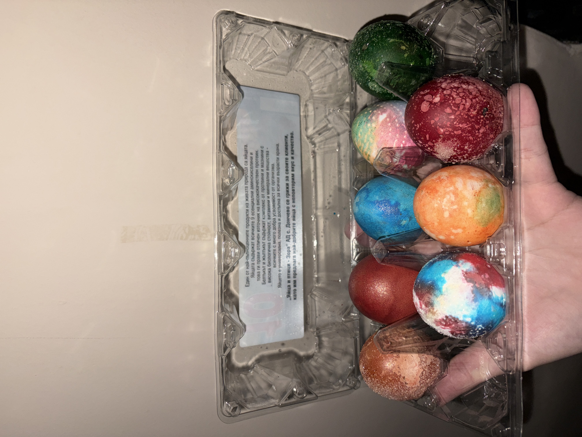 Here's a forensic description of the image:
**Subjects and Objects:** The image shows an open, clear plastic egg carton holding eight painted Easter eggs. A hand holds the carton.
**Environment:** The background consists of a light-colored wall.
**Details:**
* The Easter eggs are painted in various colors including green, red, pink, blue, orange, and brown.
* The egg carton has a label with text in Bulgarian. The label references "Яйца и птици Зора" АД с. Дончево (Eggs and Birds Zora AD c. Donchevo).
* The hand is partially visible, holding the carton.
**Location:** The label on the egg carton suggests that the eggs were manufactured by a company in Donchevo, Bulgaria. While the label is suggestive of the product origin, this information is not conclusive, and there is nothing in the image itself to conclusively establish that it was taken in Plovdiv.