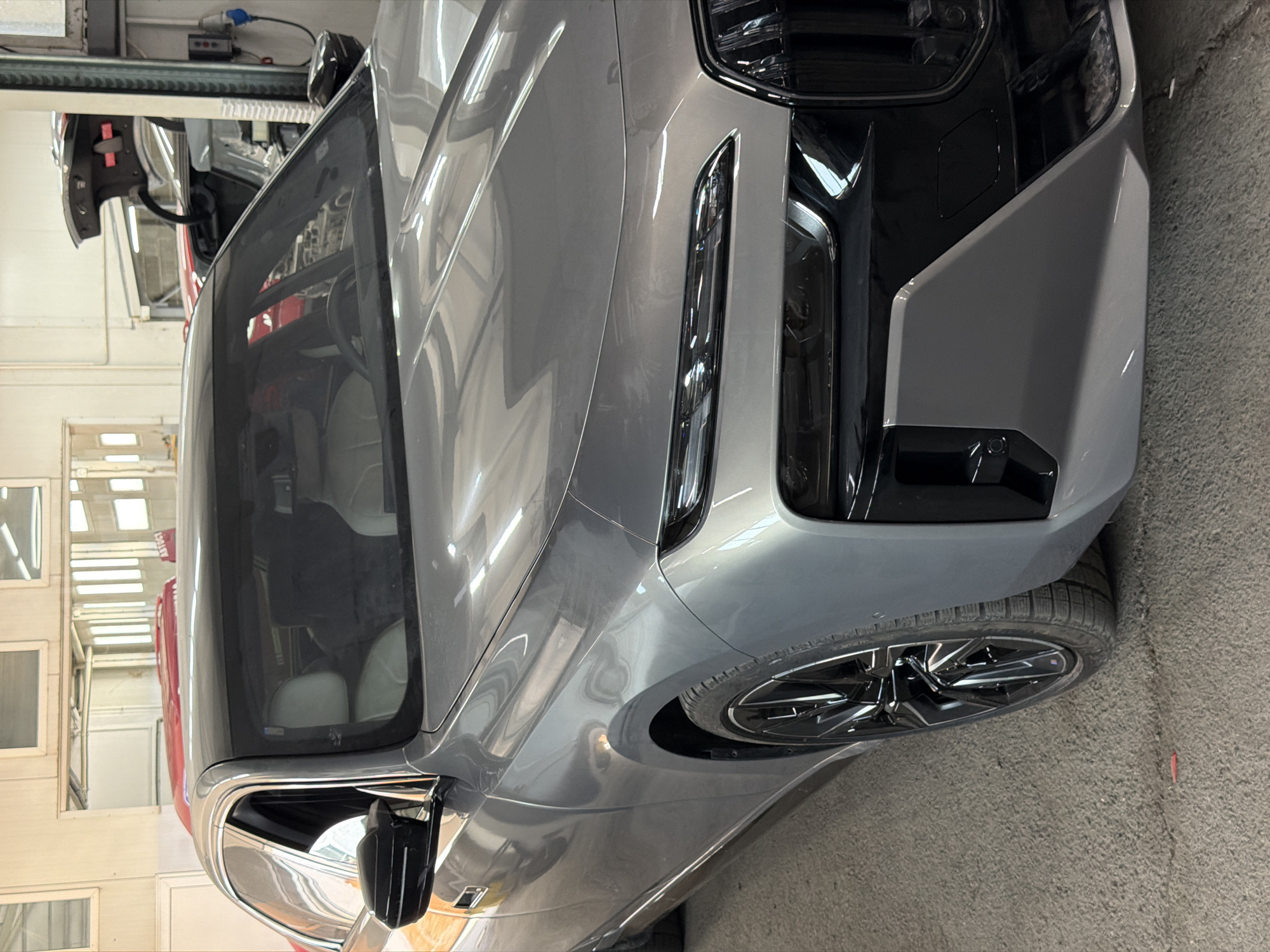 The image captures the front-left perspective of a contemporary grey luxury sedan, positioned indoors within an automotive workshop or garage. The vehicle, which appears to be a modern high-end model consistent with designs like the BMW 7 Series (G70 generation), is oriented with its front towards the right side of the frame in its corrected upright view.
The car's metallic grey finish reflects the bright interior lighting, highlighting its sleek and sculpted body lines. A distinctive feature is the vertically split headlight design, comprising upper daytime running lights and lower main headlamps. The front bumper incorporates dark, glossy black elements, contributing to a bold aesthetic. The front passenger wheel is partly visible, showcasing multi-spoke glossy black alloy rims fitted with tires. The side mirror, a combination of black and chrome, extends from the side door. Tinted windows largely obscure the interior, though light-colored seating is faintly discernible.
The indoor setting features light-colored walls and a dark, possibly concrete floor. Rectangular window-like panels or vents are visible on the left wall, reflecting ambient light. An industrial overhead structure, likely part of a vehicle lift or workshop lighting system, spans the top of the frame, fitted with a black control box and a blue power connector. A red object, potentially another vehicle or workshop equipment, is partially visible in the background on the left, bearing some indecipherable text.
The car appears stationary, suggesting it is undergoing a service, inspection, or detailing process. No individuals are present in the frame. The environment conveys a professional and well-equipped facility for vehicle maintenance, consistent with locations found in cities such as Burgas, Bulgaria.