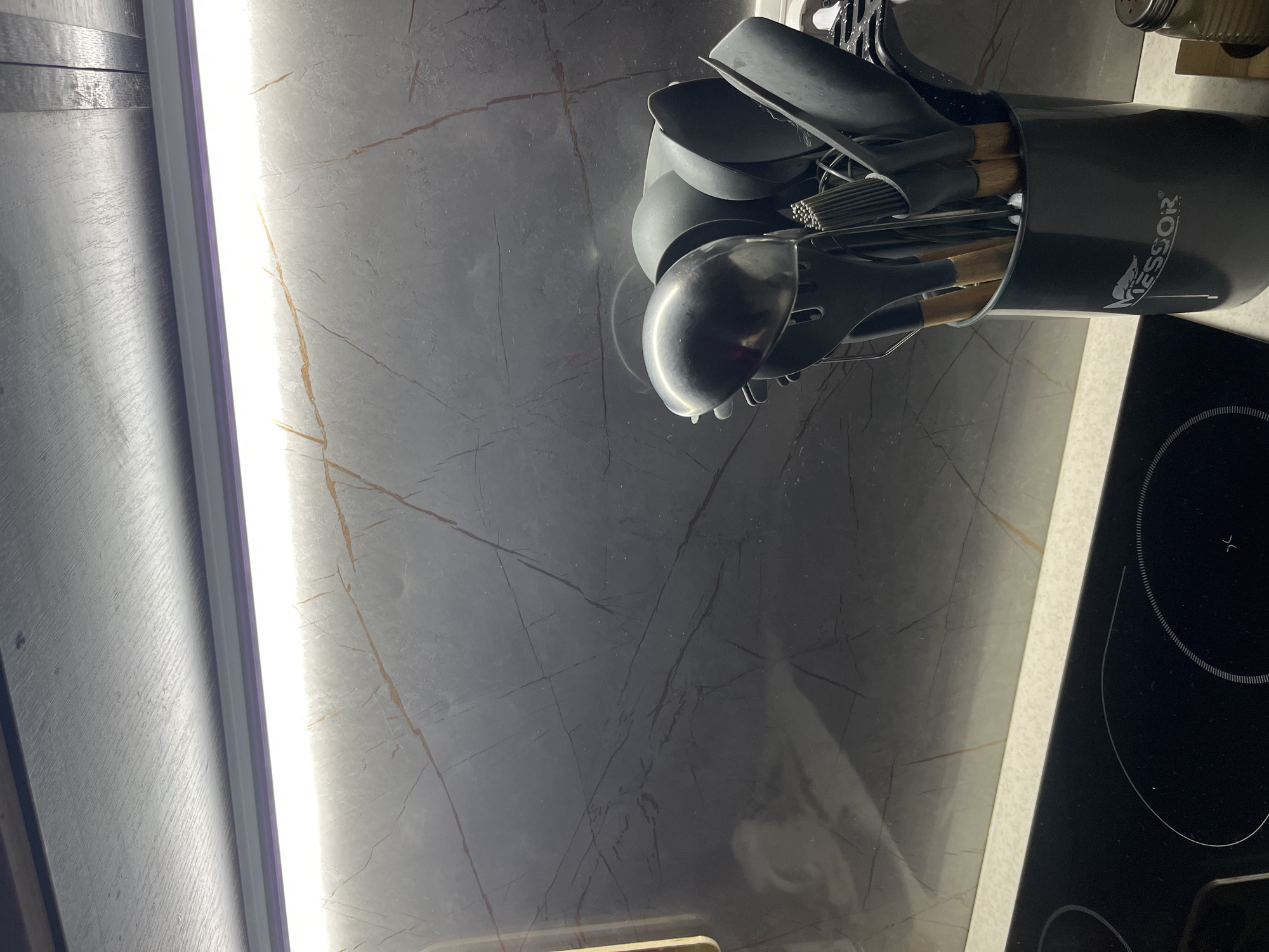 The image depicts a kitchen countertop. On the countertop, there is a black marble-patterned backsplash, an induction cooktop with two visible burners, and a black utensil holder filled with various cooking utensils, including a ladle, spatulas, and whisks. The utensil holder has the word "VESSOR" printed on it. A bright light source is visible along the left edge of the backsplash. The photograph was taken in Perm, Russia.
