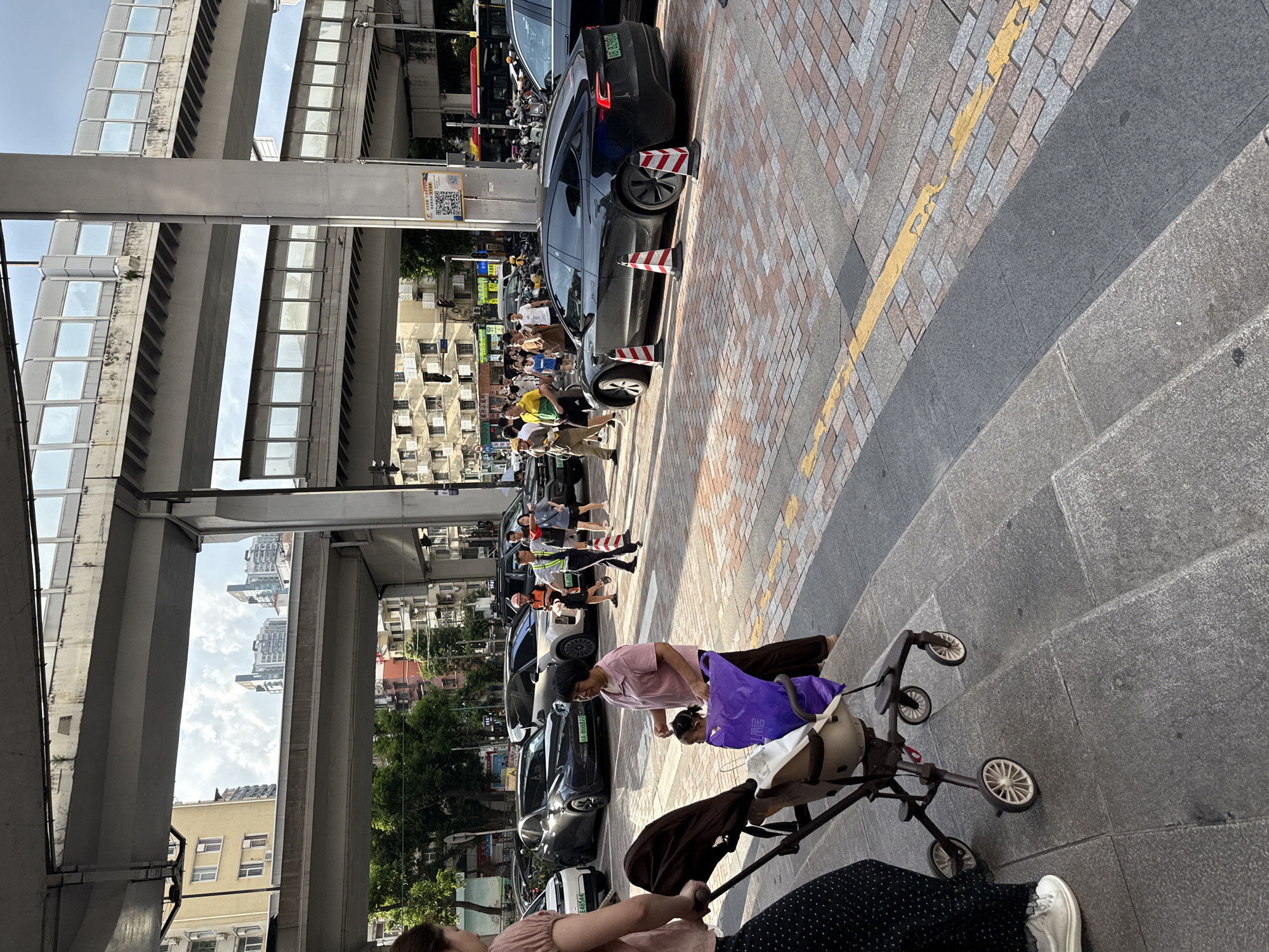 The image depicts a street scene in Haizhu District, China, under an elevated road system. In the foreground, a person pushes a stroller. To the right, an adult woman walks, holding a purple bag and looking at a child in the stroller. Several cars are parked on the street, including a black Tesla. Some traffic cones are placed around the Tesla. In the background, more buildings and elevated roadways are visible. Pedestrians are walking on the sidewalk.