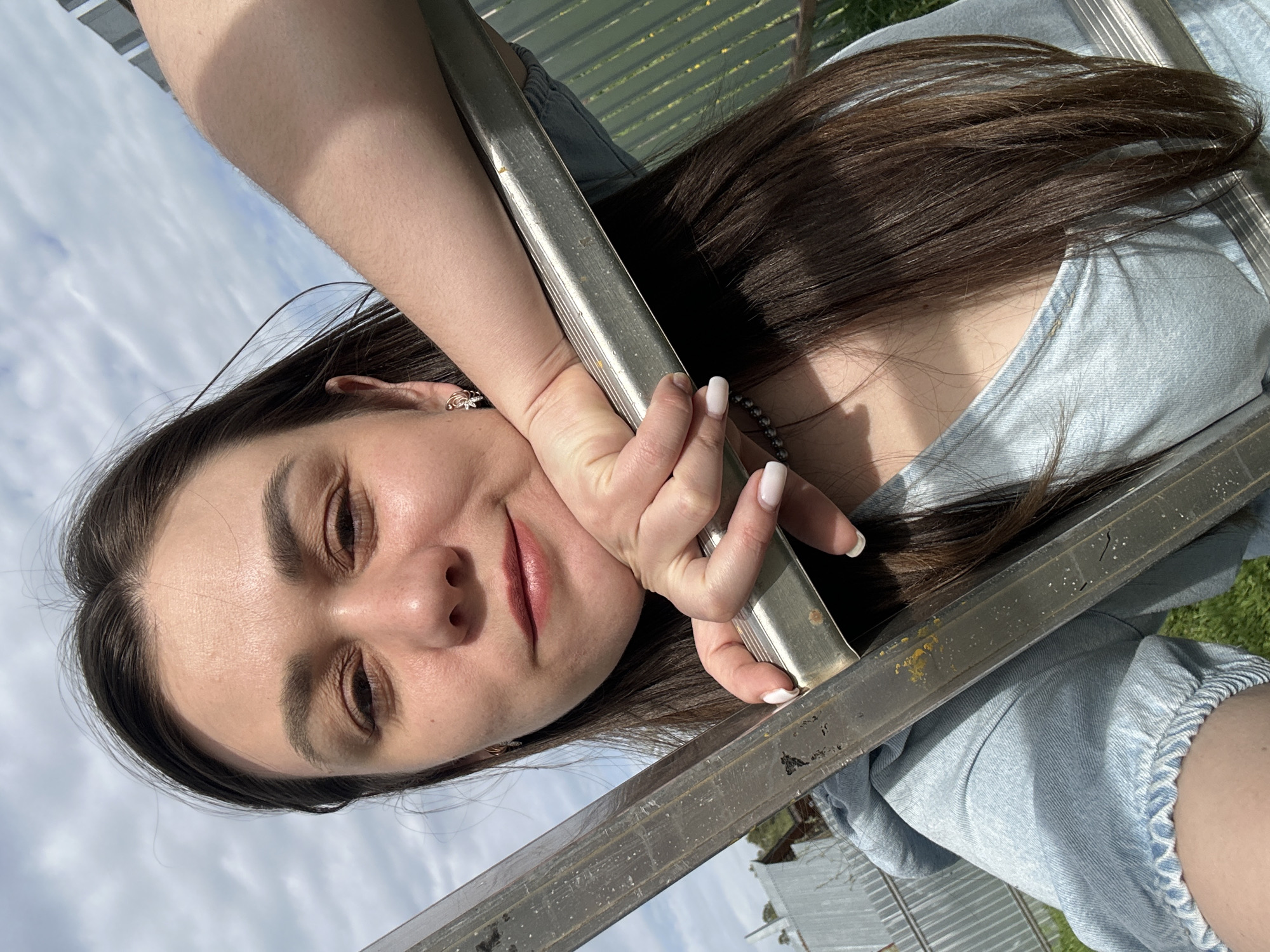 A woman is holding onto two metal bars. The woman has dark brown hair, fair skin, and is wearing a light blue denim shirt. She is holding the bars with her left hand. The background shows a sky with white clouds, a green area, and a structure with translucent panels. The photo was taken outside, and the location is Chaikovsky, Russia.