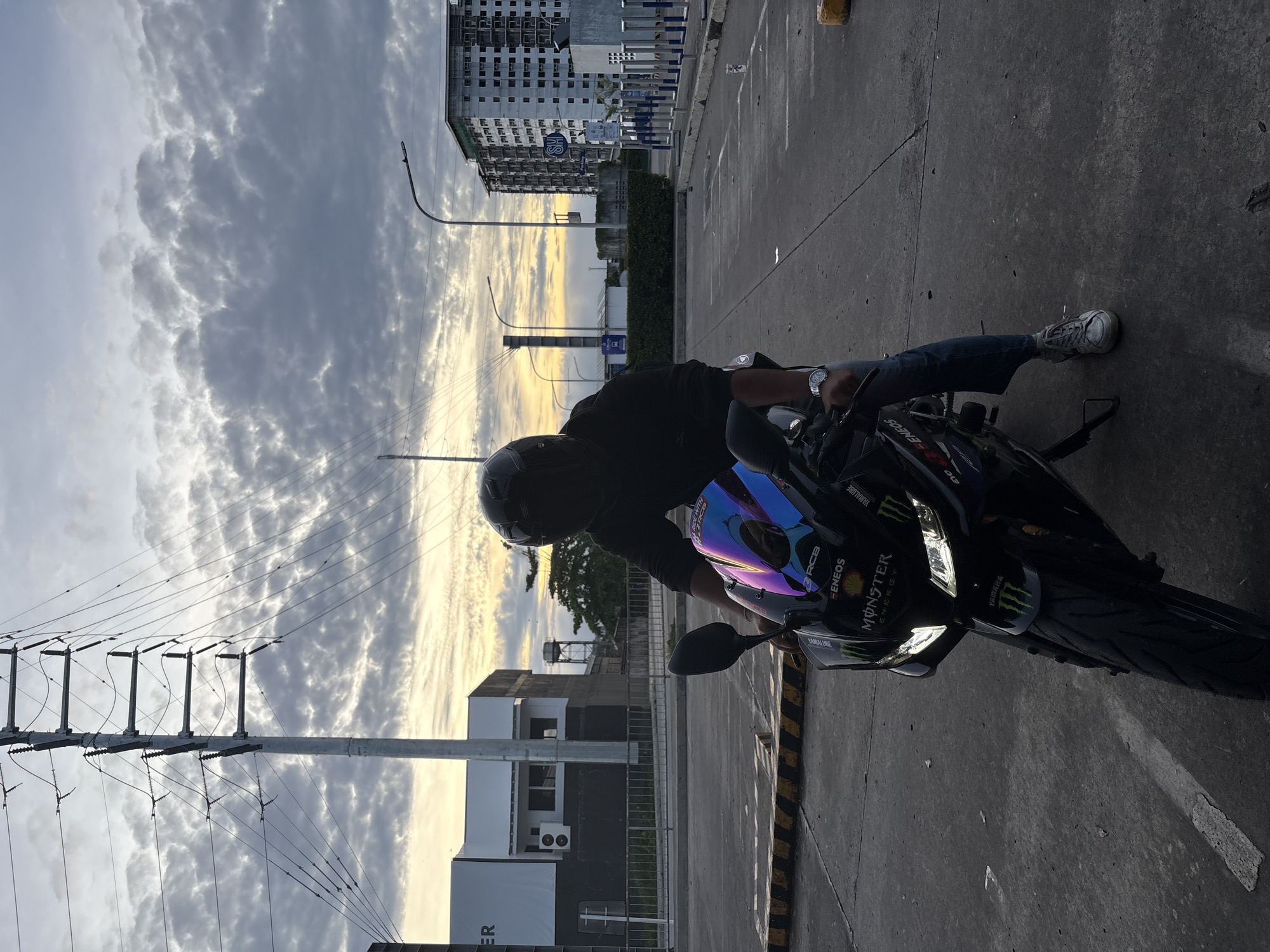 A person wearing a dark helmet and clothing is seated on a black motorcycle in what appears to be a parking lot. The motorcycle has "MONSTER ENERGY" and other decals on its front. The person is also wearing a watch on their left wrist.
In the background, there's a mix of urban elements including buildings, power lines, and street lights, as well as a cloudy sky during what seems to be sunset. The location appears to be in the city of Davao City, Philippines.