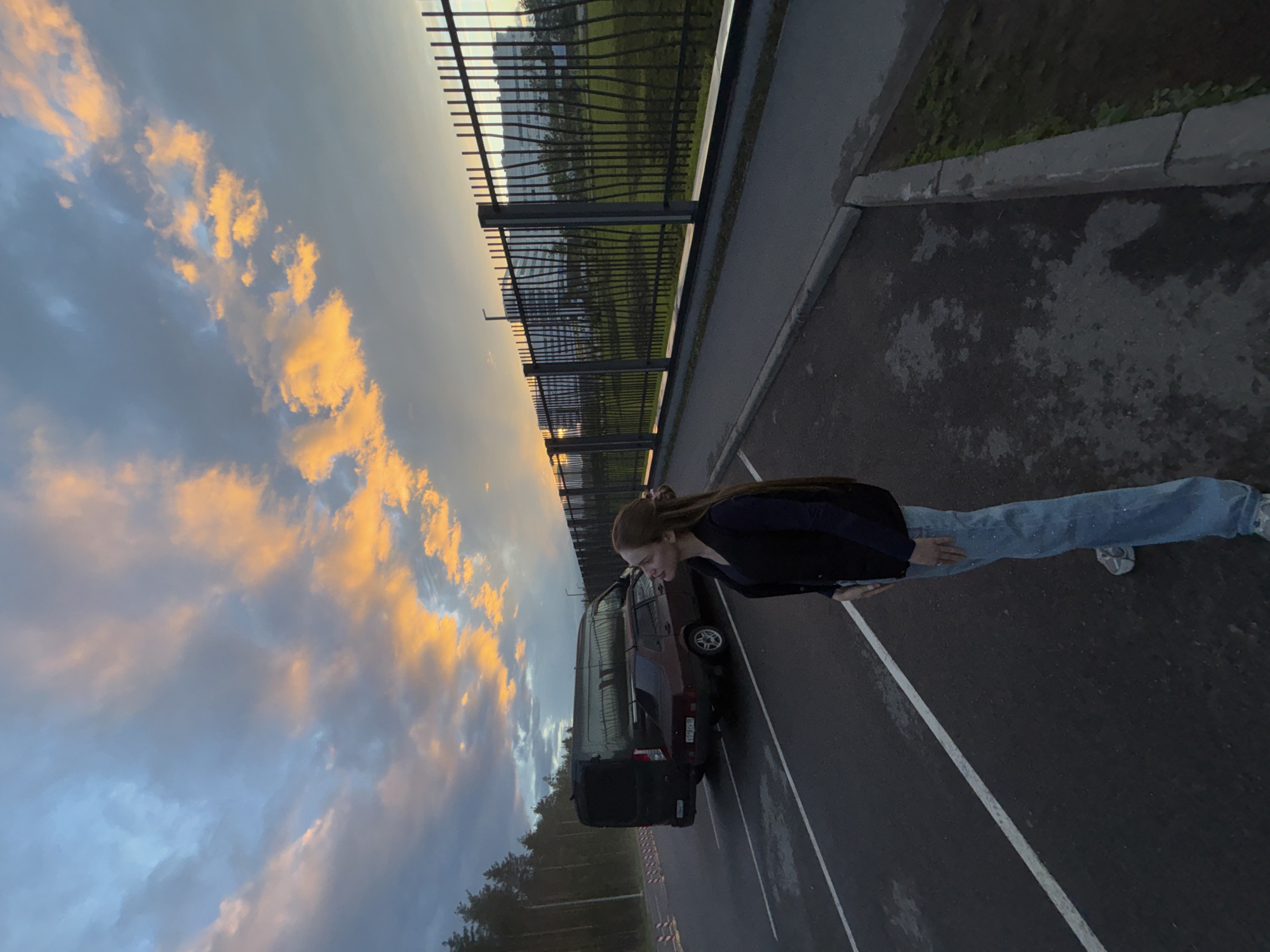 The image depicts a young woman standing in an asphalt parking lot, looking up at a dramatic sunset. The sky is filled with vibrant orange and blue clouds. Behind her, a dark-colored van and a maroon sedan are parked. A tall black metal fence with vertical bars runs along the right side of the parking lot, separating it from a grassy area and a modern building in the distance. The scene appears to be located in Arfa, Russia, indicated by the presence of a Russian license plate on the sedan. The woman is wearing a dark vest over a dark long-sleeved shirt and light blue jeans.