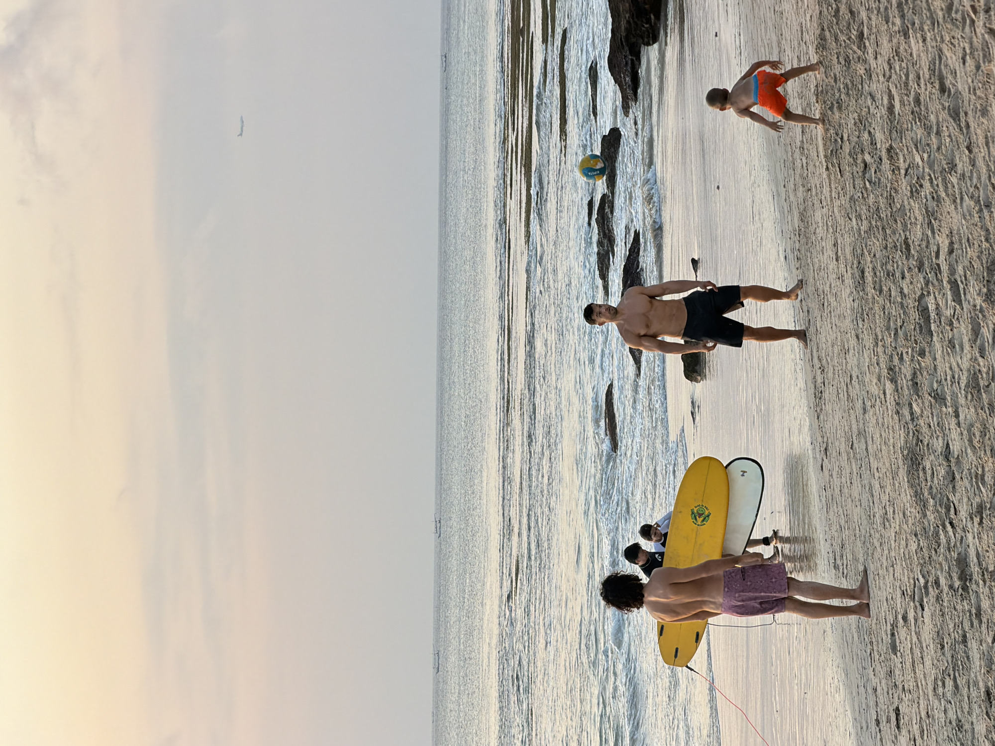 In Kuta Selatan, Indonesia, on a sandy beach, a group of people are enjoying the evening by the ocean. The sky is a soft gradient of pink and yellow, indicating either sunrise or sunset, with a faint airplane visible in the distance. The ocean has gentle waves breaking on a rocky shore.
In the foreground, a bare-chested man with dark curly hair and wearing purple patterned shorts stands facing away from the camera, holding a yellow surfboard. Behind him, two younger individuals, possibly children, are gathered around another surfboard. To the right, another bare-chested man wearing black swim trunks stands facing a young child. The child, wearing orange shorts, is crouched on the sand, looking towards the man. A colorful beach ball sits on a rock in the middle ground. Several boats are visible on the horizon, suggesting maritime activity. The scene conveys a relaxed and recreational atmosphere on the beach.