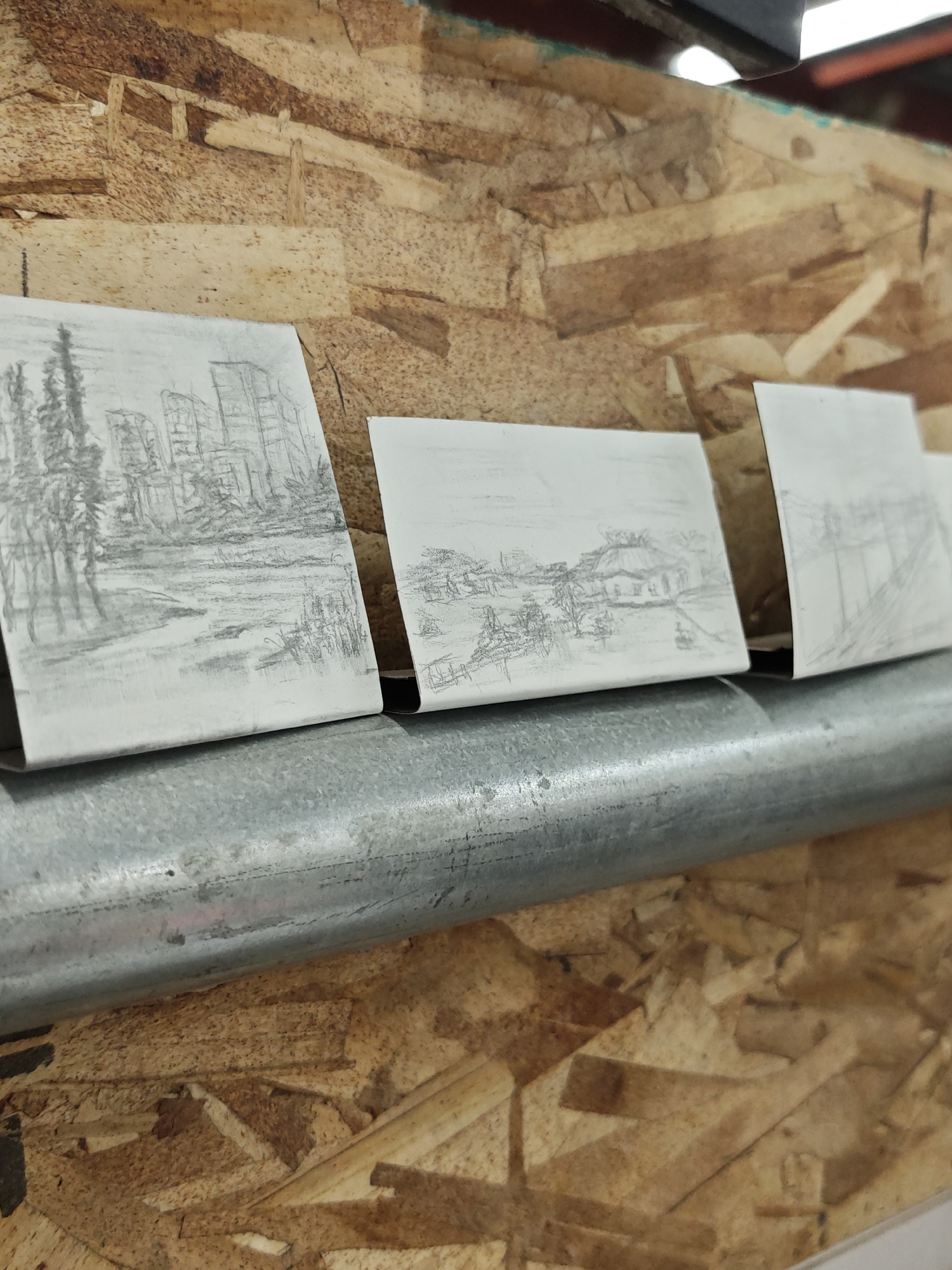 The image shows three small, white pieces of paper, each with a pencil sketch of a landscape. The papers are placed on a gray metal pipe that is mounted on a wall made of oriented strand board (OSB).
The sketch on the leftmost paper depicts a scene with tall, thin trees in the foreground, and what appear to be buildings in the background. The style is impressionistic, with rough lines and shading. A body of water, possibly a river or lake, is also suggested in the foreground, with reflections of the trees and buildings.
The middle sketch shows a rural scene, with a building that resembles a house or barn with a pitched roof. There are trees and possibly some fences or other structures around it. The sketch is less detailed than the first one.
The rightmost sketch is the most abstract of the three, with only faint lines suggesting some structures, possibly utility poles or fence posts.
The OSB wall has a rough, textured surface with visible wood chips and variations in color, ranging from light beige to darker brown. The metal pipe is cylindrical and appears to be industrial in nature, with a slightly weathered surface. The overall lighting suggests an indoor environment, possibly a workshop or storage area.
There is no identifiable information within the image to suggest a specific location in Calgary, Canada. The sketches themselves are generic landscapes and do not contain any recognizable landmarks or cultural elements specific to any city.
