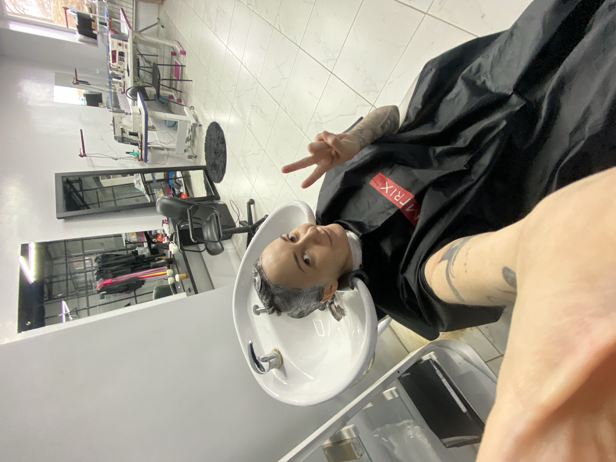 A person with short, gray hair is sitting in a salon chair with their head tilted back into a white wash basin. They are wearing a black salon cape with the word "MATRIX" printed on it. The person is making a peace sign with their right hand. Their arms are visibly tattooed. The background shows a tiled floor, mirrors, and salon equipment, indicating the person is at a salon. The exact location is not possible to determine with only the given information.