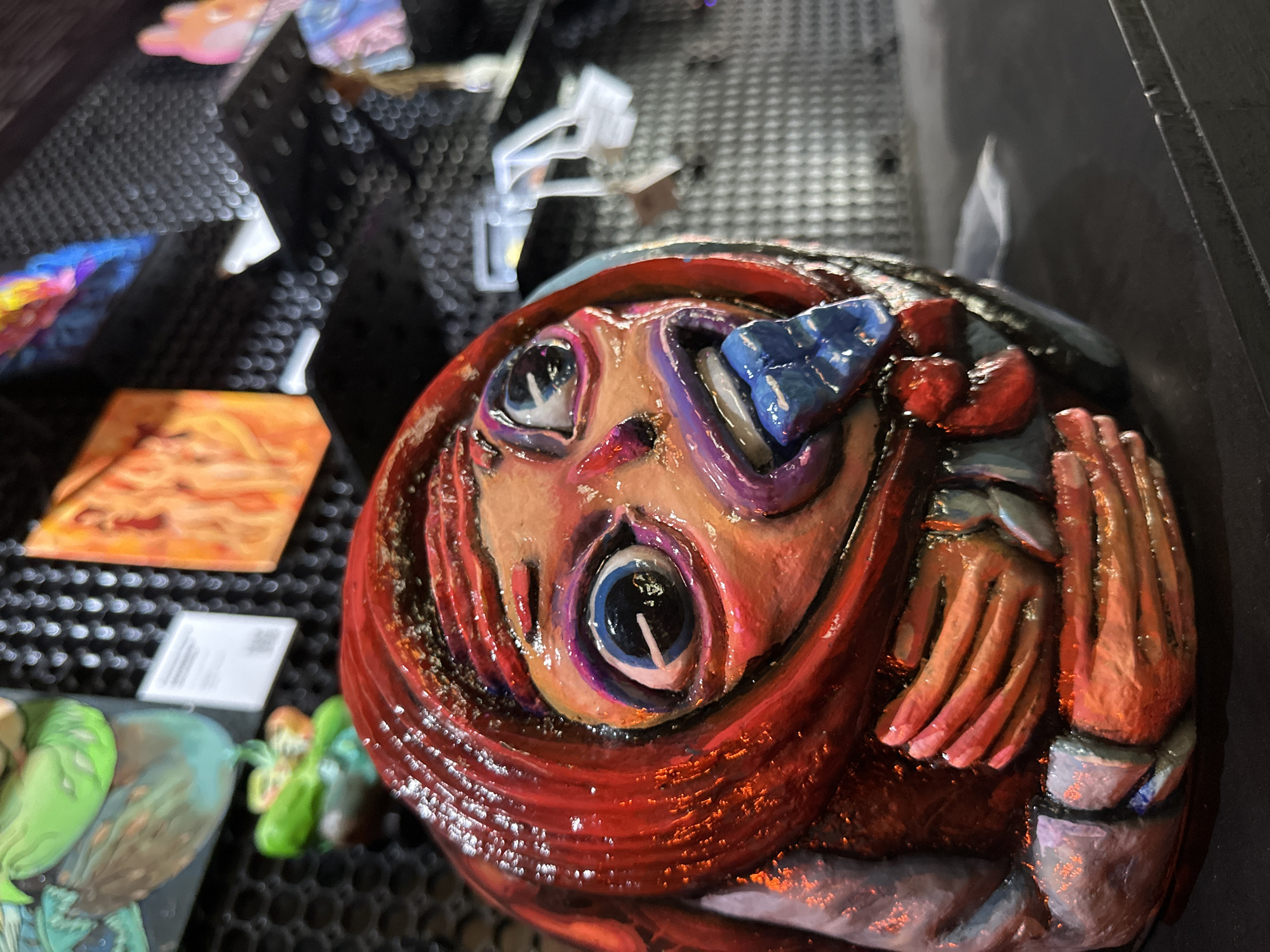 The image shows a close-up of a unique, possibly handmade, art piece. The piece depicts a distorted human face with exaggerated features. The face is framed by what appears to be a red, textured material that could represent hair or a garment. The skin is painted in varying shades of orange and pink, with purple accents around the eyes. One eye socket is filled with a large, blue, geometric object. To the side of the face are painted hands. The artwork has a glossy finish, suggesting a resin or varnish coating. The artwork is displayed on a black, perforated metal surface alongside other small artworks or objects. The background is dark and slightly blurred. The location ตำบลเกาะพะงัน, Thailand is only speculative as there is nothing in the image that directly identifies it as being located there.
