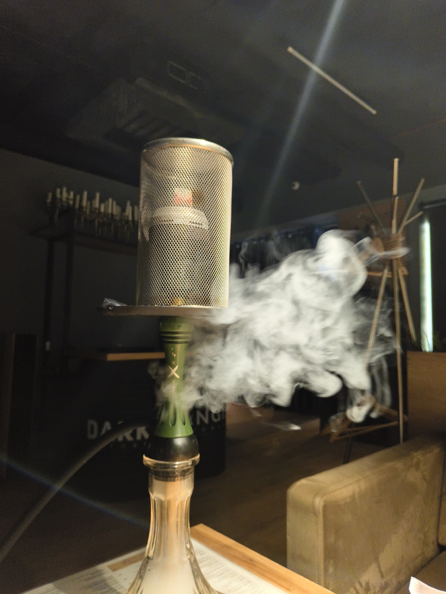 A still, indoor shot of a smoking hookah in a dimly lit environment in the city of Unknown, Unknown. The hookah, with a clear base and green stem, sits on a wooden surface, presumably a table. It is topped with a metal mesh cage containing a red heating element. Dense white smoke billows out from around the hookah, partially obscuring objects in the background. Behind the hookah, partially out of focus, there are shelves with glassware, dark containers labeled "DARK", and a structure of wooden sticks. A neutral-colored sofa is visible on the right side of the frame. The ceiling has a dark hue, with a light fixture hanging from it.