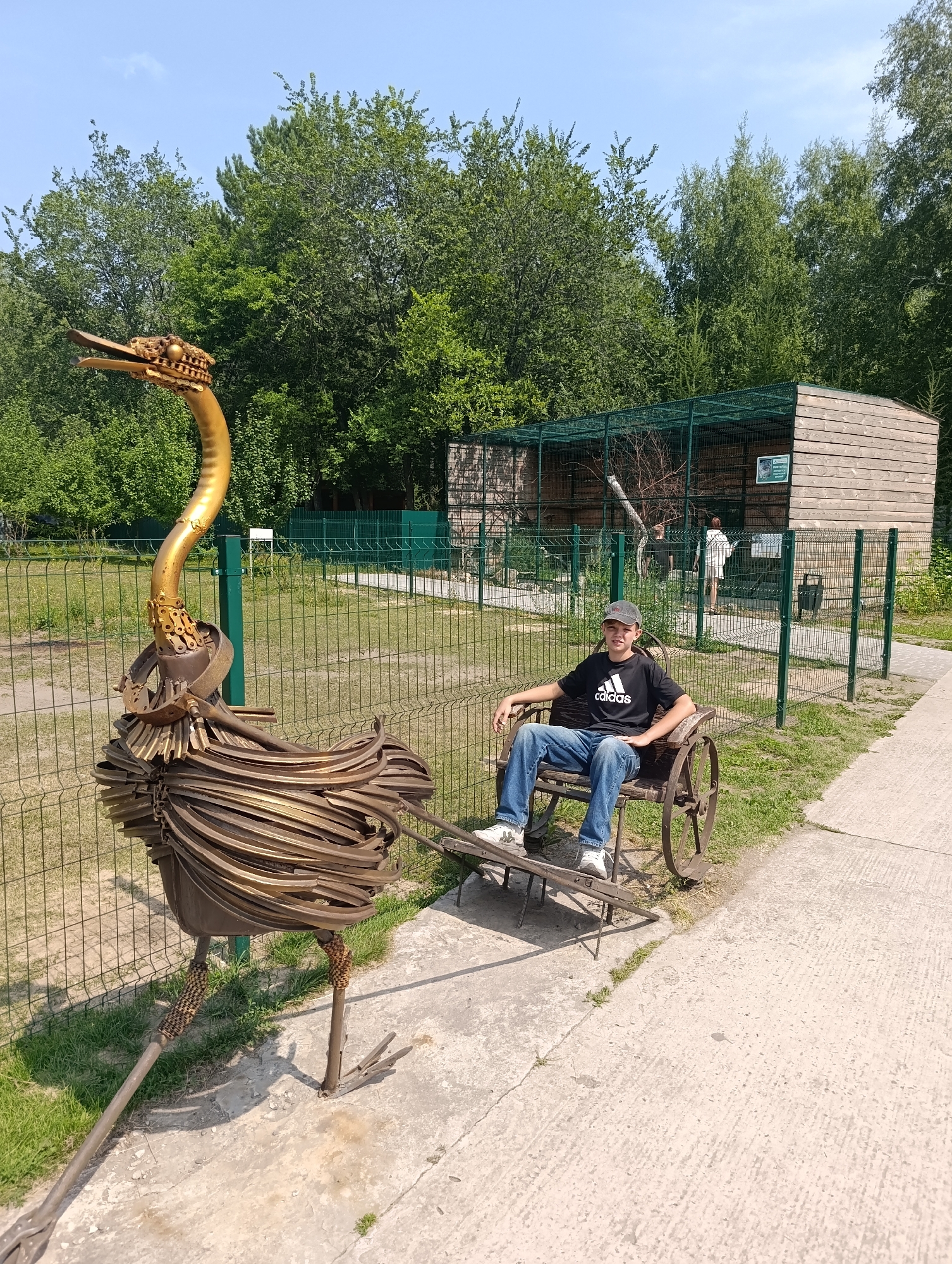 A young male with dark hair, wearing a grey baseball cap, black t-shirt with white Adidas logo, blue jeans, and white sneakers, is sitting in a rustic wooden cart with spoked wheels. He is positioned in the foreground, facing forward. To his left, a large, intricate metal sculpture of an ostrich stands. The sculpture is predominantly gold and brown, with a long neck and body constructed from layered metal elements and chains. It has a beak-like head with visible metal detailing and two prominent legs with metal feet. The cart and ostrich are situated on a concrete pathway.
In the background, a fenced enclosure with a wooden structure and wire mesh is visible. This enclosure appears to be an animal habitat, possibly at a zoo or park. Through the mesh, two individuals can be seen, one standing and another partially obscured. A metal fence with vertical posts and horizontal wires runs across the middle ground, separating the pathway from a grassy area and the enclosure. Lush green trees form a dense backdrop to the scene under a clear blue sky with a few scattered clouds. The overall environment suggests an outdoor recreational area during daytime.