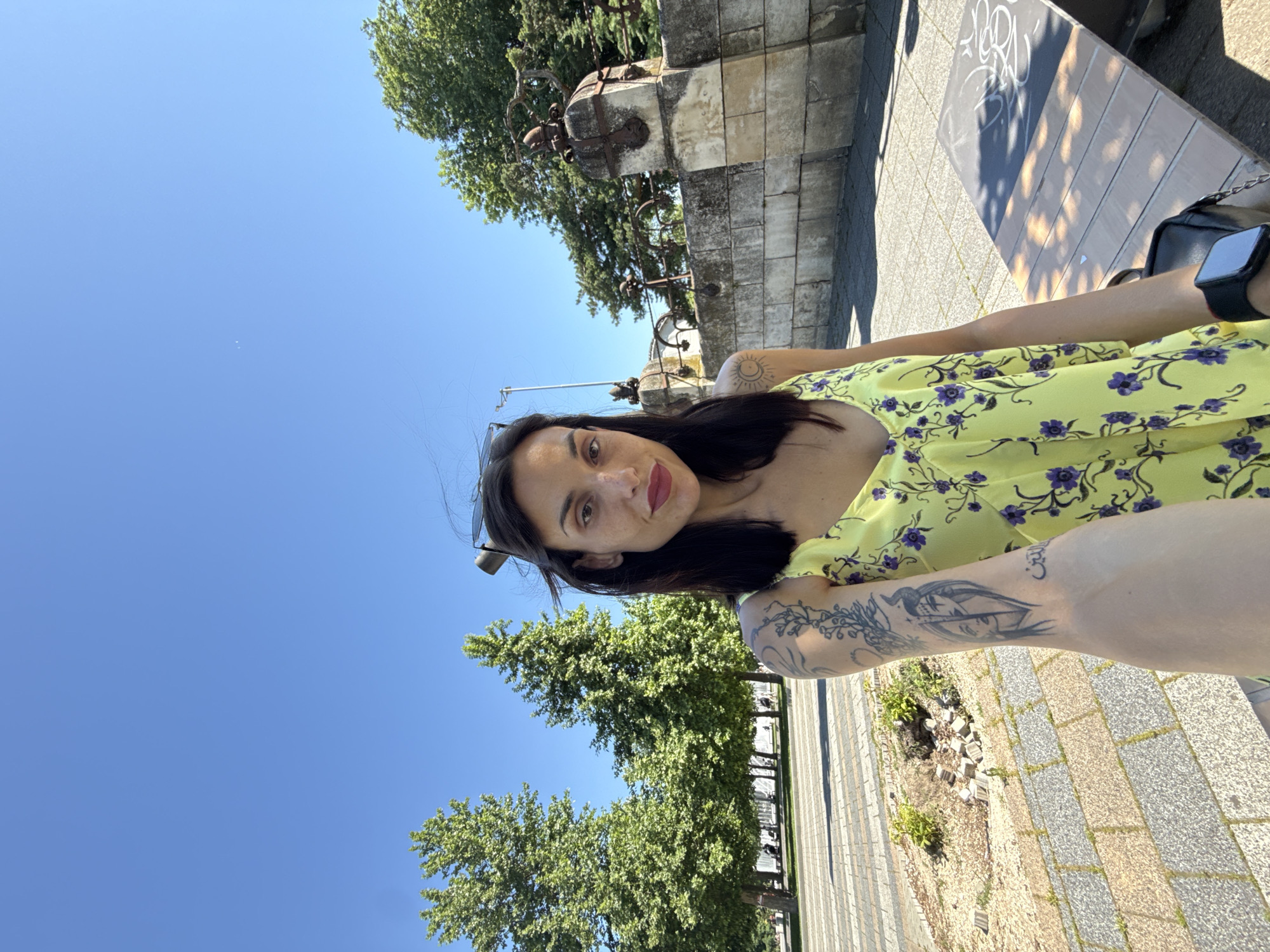 A woman is posing for a selfie outdoors, likely in Pleven, Bulgaria, considering the architecture and environment. She has dark hair and olive skin, and is wearing a yellow floral dress. The woman has visible tattoos on her arms and shoulder. She appears to be seated on a bench next to a paved walkway. Behind her, there is a stone wall with decorative metal railings and lush green trees, set against a clear blue sky. The scene appears to be taken during daytime.