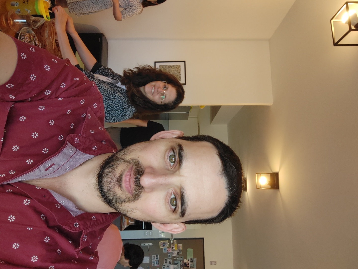 This indoor image, likely taken at a social gathering, features three individuals and is presented rotated 90 degrees counter-clockwise. When viewed in its intended upright orientation, the scene appears to be an evening event, indicated by the prominent artificial lighting.
In the foreground, a man with dark hair, a beard, and green eyes looks directly at the camera with a neutral expression. He is wearing a maroon button-up shirt adorned with small white floral patterns.
Behind him, in the mid-ground, a woman with long, curly brown hair and glasses smiles towards the camera. She is dressed in a patterned top, possibly a leopard print, worn over a dark t-shirt. Her hands are visible, resting on a surface.
Further in the background, to the right of the smiling woman, another person is partially visible, wearing a light-colored, patterned dress or shirt. In the lower left background, the back of another person's head is visible near a bulletin board covered with various pinned items.
The setting appears to be a modern home or an informal event space in Tel-Aviv, Israel. A table in the mid-ground holds a clear glass containing a dark liquid and a yellow plastic cup. The light-colored walls are illuminated by the warm glow of two wall-mounted sconces: a hexagonal one in the upper right and a simpler one positioned centrally on the right wall. A framed picture hangs on the wall behind the smiling woman. The overall atmosphere suggests a relaxed and convivial event. There is no visible text in the image.