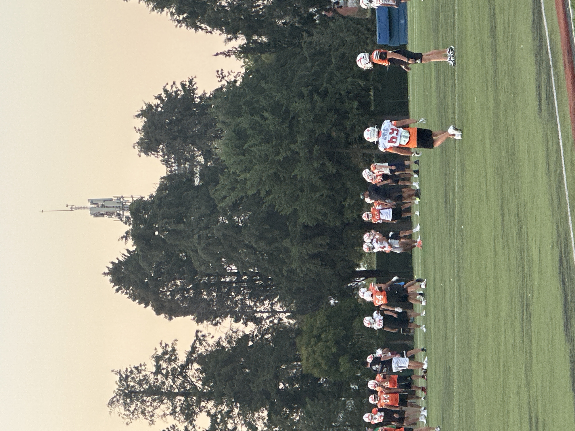 An outdoor American football practice or training session is captured on a clear day, likely in the late afternoon or early evening due to the soft, pale yellow hue of the sky. Numerous individuals, primarily male, are visible on a green grassy field marked with white lines, with a red track lane also present on the right side.
The participants are dressed in American football practice attire, including helmets, jerseys, shorts, and cleats. The jerseys display a mix of colors: white with orange numbering and details, orange with white, and some black. Several players' white jerseys clearly show the text "AZTECAS." Many of the players are standing in lines or grouped together across the field, suggesting organized drills, warm-ups, or instructions. One player, prominently featured, wears a white jersey with the number "59" and "AZTECAS" text.
In the background, dense green trees create a natural boundary, beyond which a tall communications tower with multiple platforms and antennas rises against the sky. Some blue practice pads are also discernible behind the players. The scene depicts a focused athletic environment, typical of a sports training facility in San Andrés Cholula, Mexico.