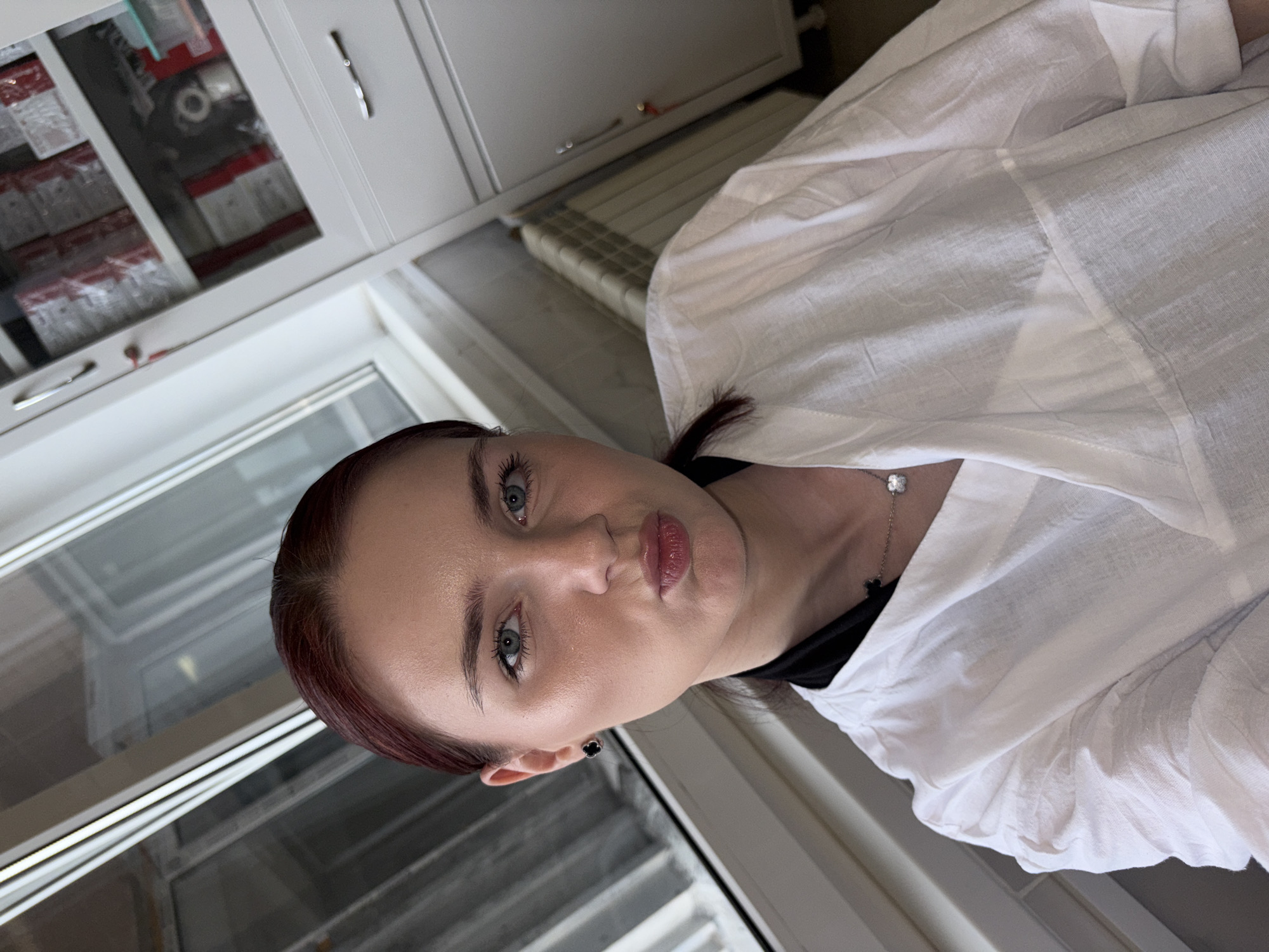 A woman with auburn hair and blue eyes is pictured in a close-up shot, angled upwards. She is wearing a white garment over a black top, with a silver necklace featuring a clover-shaped pendant visible. Her makeup includes mascara, eyeliner, and lip gloss. The background shows a white cabinet with a glass door containing shelves of items, a radiator, and a window. The scene appears to be indoors, likely in a domestic or professional setting, with no specific geographical markers of поселок №13, Russia.