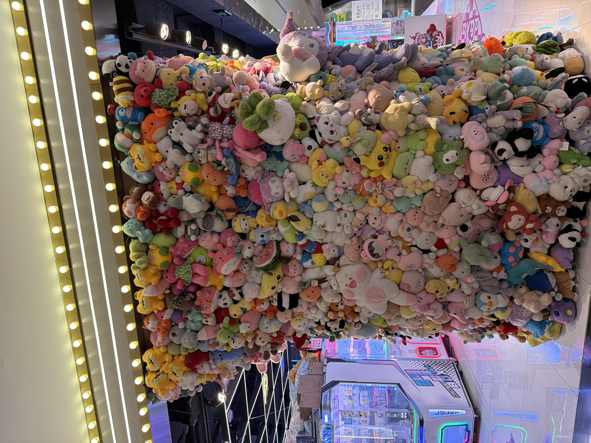 This image captures a vibrant, vertically oriented scene dominated by a colossal pile of colorful plush toys. Thousands of stuffed animals, ranging from small to medium-large, are densely packed together, forming what appears to be an elaborate display or a massive collection within an entertainment venue. The plushies depict a variety of popular characters and animals, including pandas, cats, bears, and known figures like Pikachu and My Melody, in a diverse palette of pastel and bright colors.
The setting is an indoor space, likely an arcade or a specialized toy store, suggested by the presence of game machines in the lower right portion of the frame. One prominent feature on the left is a vertical structure adorned with strips of warm yellow bulbs and bright white fluorescent lights, providing ample artificial illumination to the scene. The overall impression is one of abundance and playful excitement.
Visible text in the image includes: "LUCKY", "WELCO" (likely part of "WELCOME"), "CRAZY BOXES" on an arcade machine, "JOYOUS", "FREE LUCK", and Chinese characters "包店员" (bao diànyuán, potentially "shop assistant" or related to packaging) and "免费咨询" (miǎn fèi zī xún, meaning "free consultation"). The numbers "2" and "12." are also partially visible.