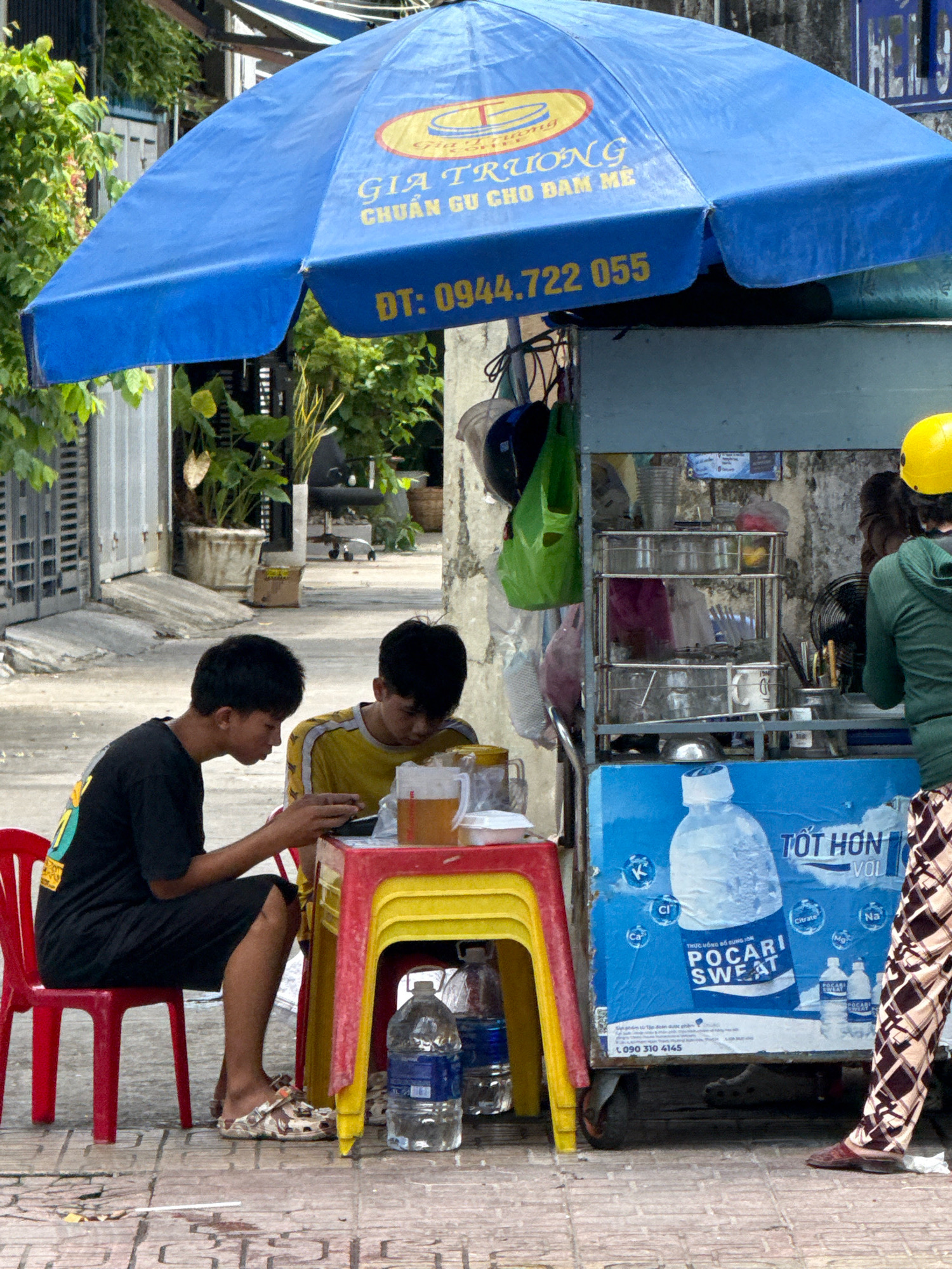 The image depicts an outdoor street scene in Nha Trang, Vietnam, featuring a food and beverage stall under a large blue umbrella. Two young males, likely teenagers, are seated at a red and yellow plastic table. The male on the left wears a black t-shirt and dark shorts, while the one on the right has on a yellow t-shirt with yellow stripes. They appear to be engaged with a mobile device and drinking from glasses containing a light-colored liquid, possibly tea. The stall itself is equipped with shelves displaying various items, including plastic cups, bottles, and what appear to be food containers. A yellow helmet is visible near the stall, suggesting a person on a motorcycle may be nearby. The ground is paved with square tiles, and a red plastic chair is positioned to the left of the table. The background shows a building facade and lush green vegetation. A large poster for "POCARI SWEAT" is affixed to the side of the stall. The overall atmosphere is casual and everyday, characteristic of a street-side vendor in Vietnam.