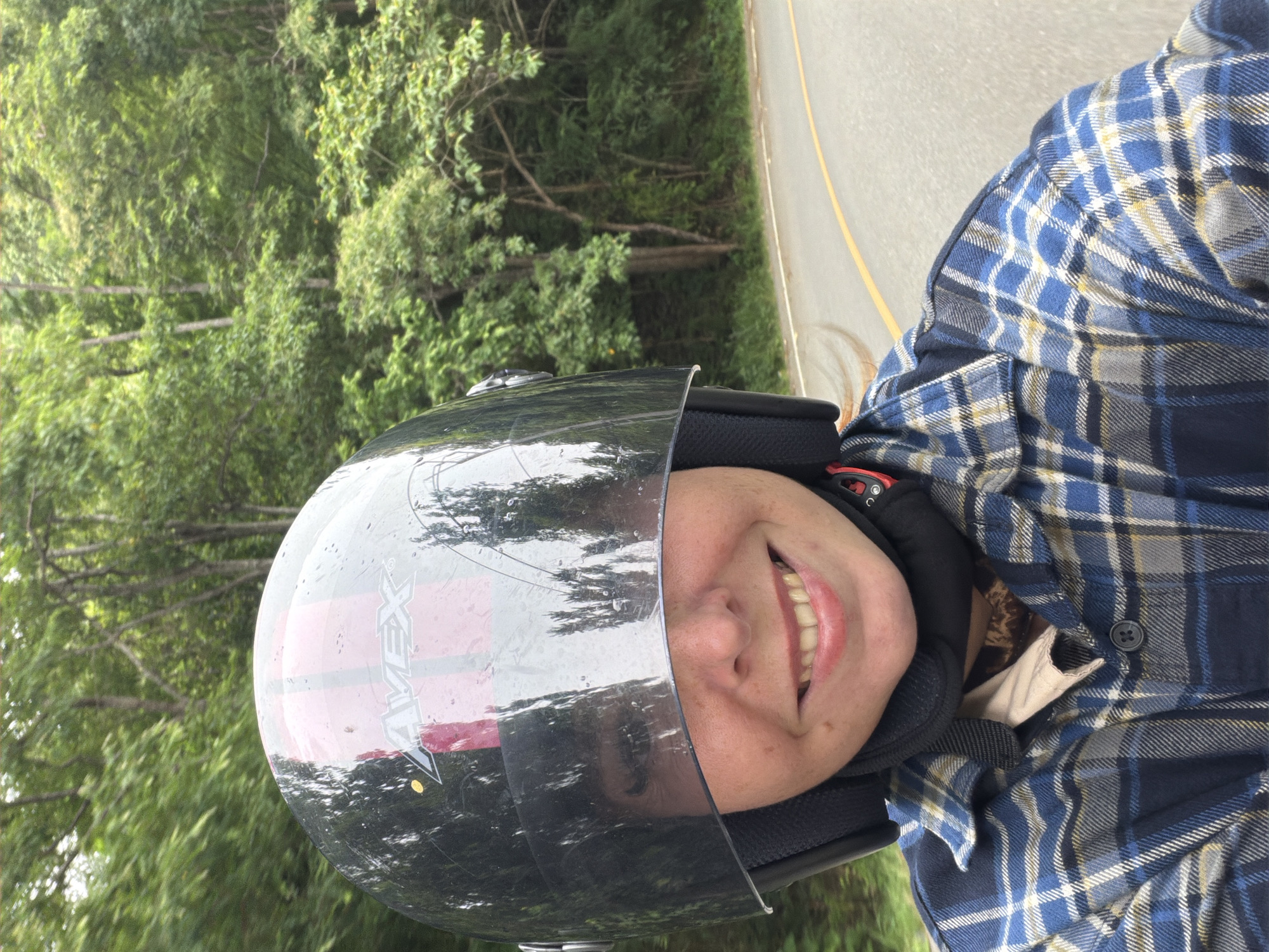 In Samoeng Tai, Thailand, a person wearing a black and pink helmet with a reflective visor is captured in a selfie. The visor reflects trees and the sky, obscuring the upper part of their face. The person is smiling broadly, revealing their teeth. They are wearing a blue and white plaid shirt over a tan garment. Their hair, light brown, is visible on the side, and a black neck protector is worn. The background shows a paved road curving through a lush, green forest with tall trees. The road has a yellow line marking the edge. The image appears to be taken from a moving vehicle, likely a motorcycle, given the perspective and the person's attire.