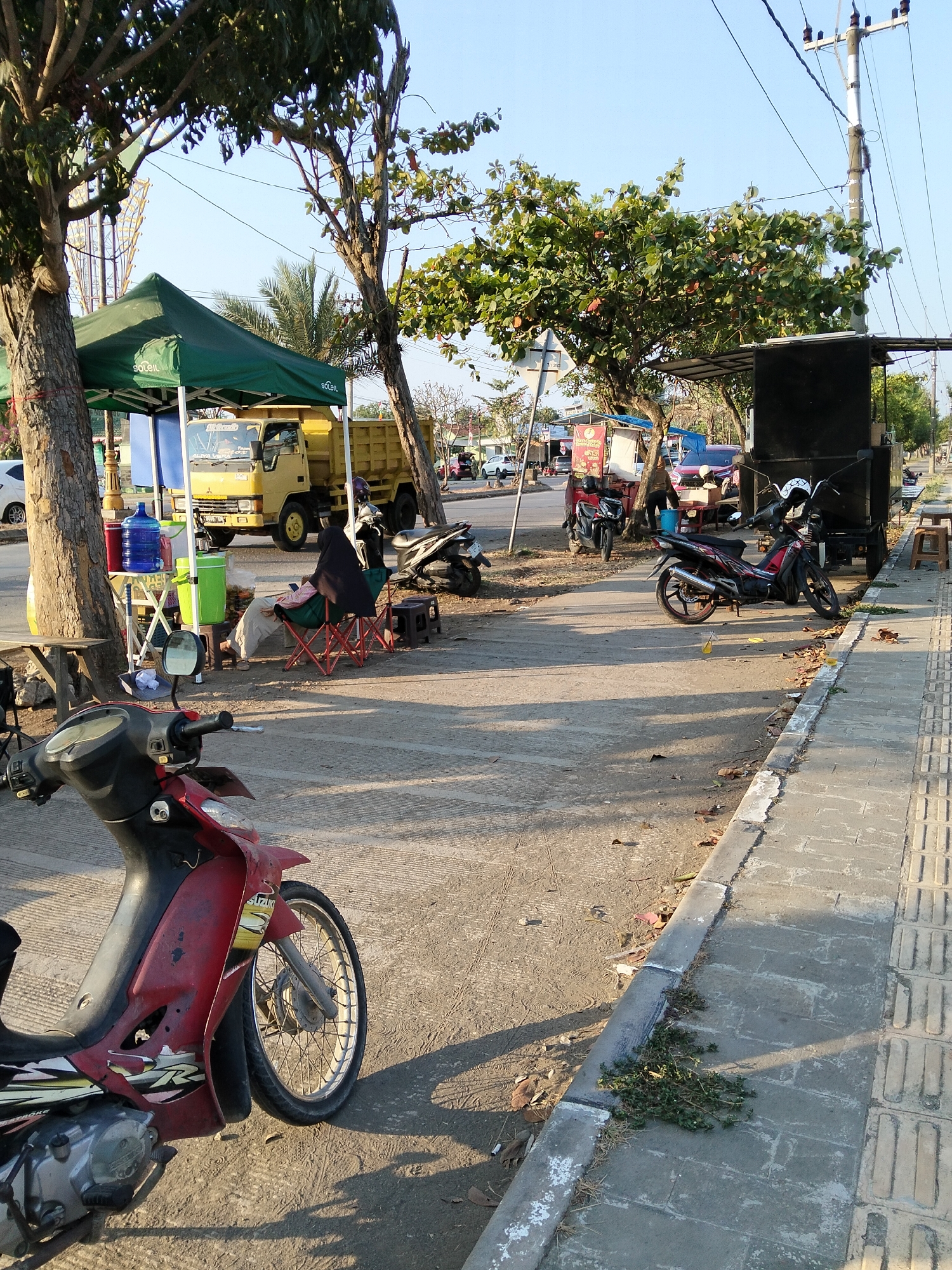 The image depicts a street scene in Indramayu, Indonesia, likely during the late afternoon due to the long shadows. The foreground features the front of a red and black motorcycle. In the midground, a green tent covers a stall or vendor area, with a yellow dump truck parked behind it. Several water containers, including blue and green dispensers, are visible at the stall. A person wearing a black head covering and light-colored clothing is seated in a red folding chair near the stall. Further to the right, other motorcycles are parked along the sidewalk. The background shows a street with some traffic and buildings. The environment is characterized by a paved road, a concrete sidewalk with tactile paving, trees lining the street, and a clear sky. Power lines and a utility pole are also prominent. The overall impression is of a busy, utilitarian streetscape in a tropical or subtropical urban setting.