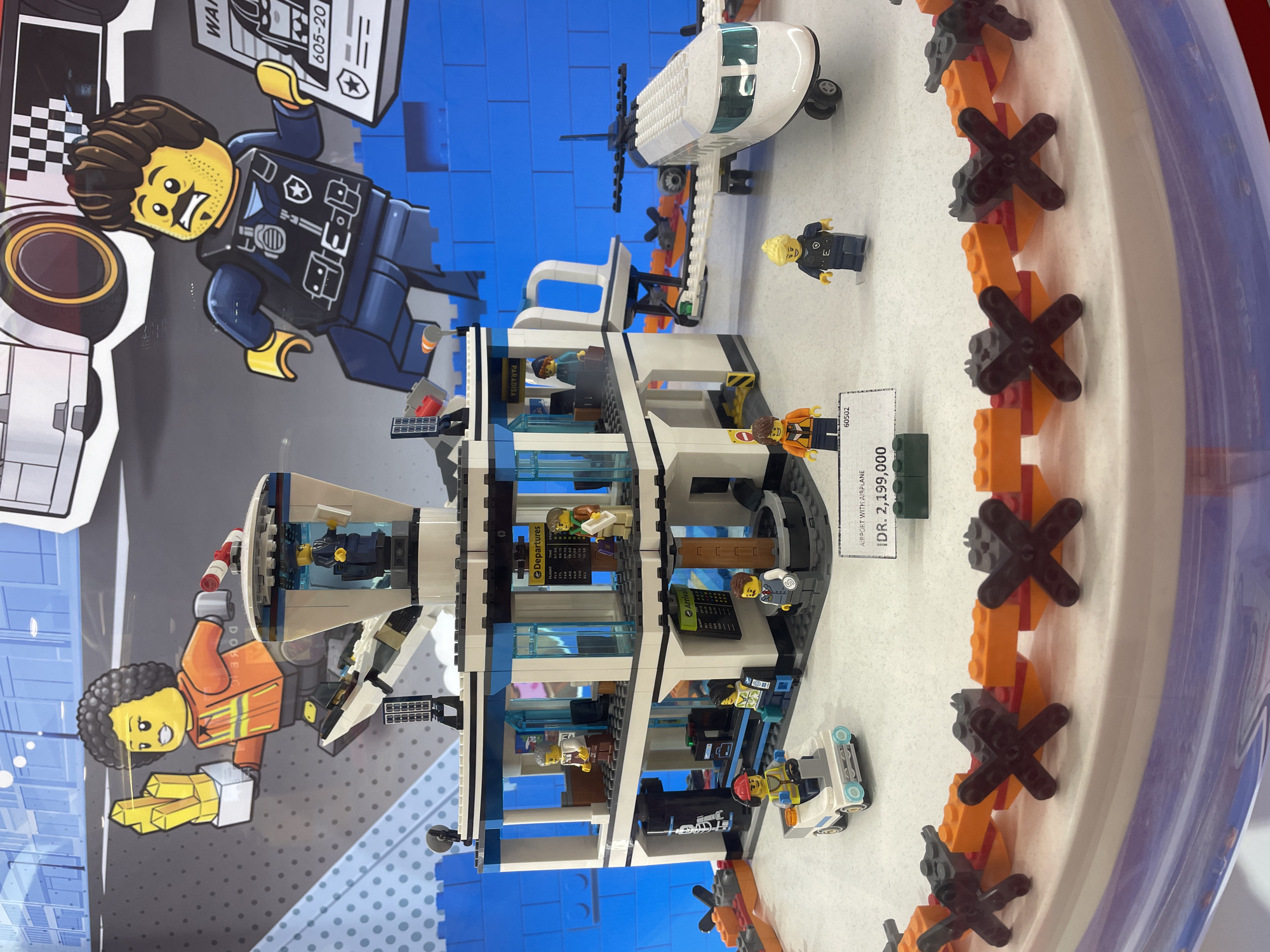 The image displays a Lego airport set, likely part of a retail or exhibition display in Denpasar, Indonesia. The central subject is a multi-story Lego airport building, bustling with numerous Lego minifigures engaged in various activities, such as checking in at "Departures," waiting, and staff working at service counters.
Outside the airport building, a white Lego helicopter with a black rotor is positioned on the right, and a small white Lego service vehicle with a minifigure driver is near the front. The display features a decorative border of orange and black "X"-shaped Lego pieces, possibly resembling runway markers.
Flanking the main Lego set are large, vibrant cardboard cutouts of two Lego characters. On the left, a Lego police officer with a mustache and goatee smiles, holding a grey box with text that partially reads "WAN" and "605-20." On the bottom left, another Lego character with dark curly hair, wearing an orange jumpsuit, smiles and points.
Visible text on the airport building includes "Departures," "Arrival," and "PARADISA." A price tag in front of the set clearly states "AIRPORT WITH AIRPLANE" and "IDR. 2,199,000," which is the price in Indonesian Rupiah, confirming the location context of Denpasar, Indonesia. The set number "60502" is also visible on the tag. The image is captured indoors under artificial lighting.