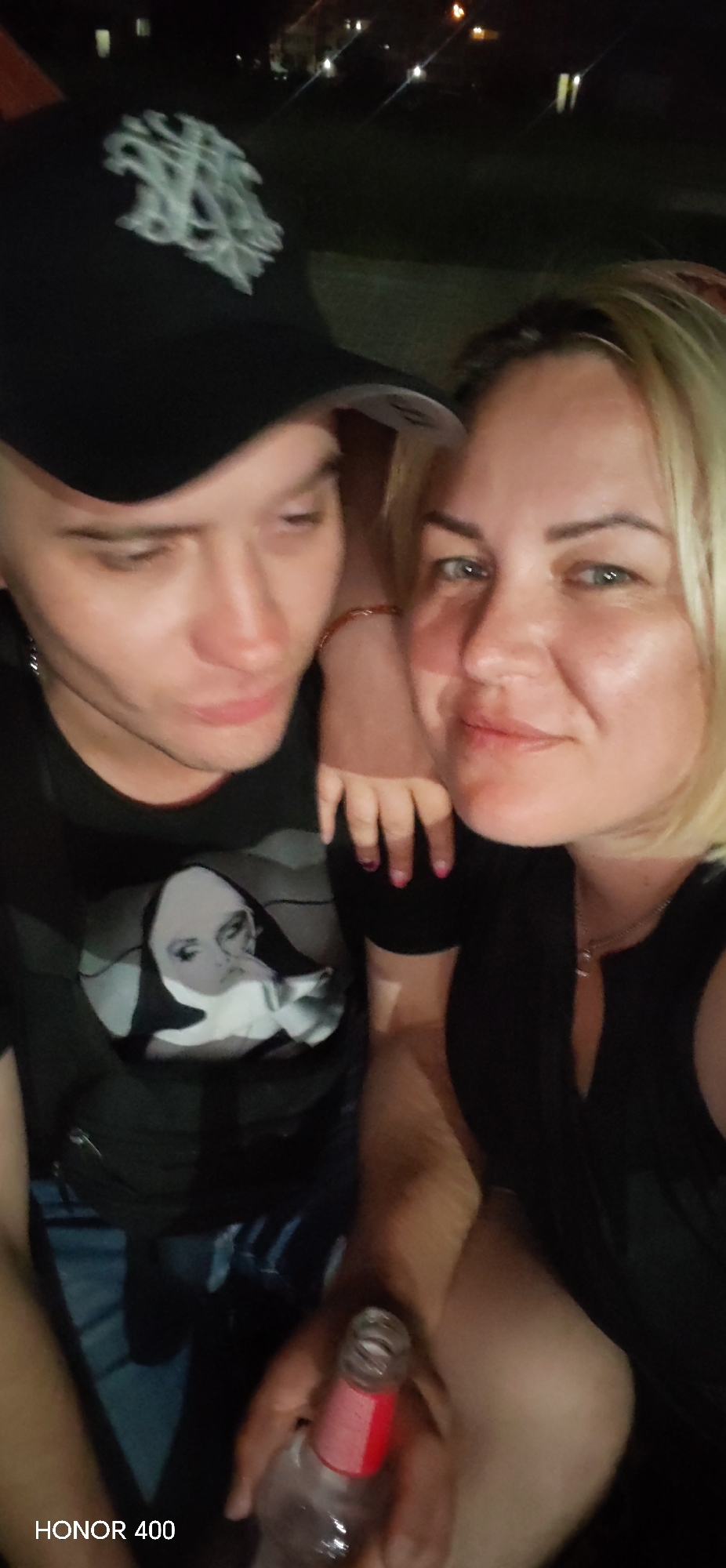 In this close-up, low-light selfie, two individuals are positioned in the foreground. On the left, a person wearing a black baseball cap with a white emblem and a black t-shirt featuring a graphic of a stylized face is partially visible. Their face is turned to the side, looking downwards. To the right, a woman with blonde hair and light-colored eyes is looking towards the camera. She is wearing a black top and a necklace. Her right arm is resting on the person on the left's shoulder, and her hand is visible with pink nail polish. In the lower right of the frame, a partially consumed bottle with a red label is held by an unseen hand. The background is dark and out of focus, with distant lights suggesting an urban or suburban setting at night. The text "HONOR 400" is visible in the bottom left corner of the image. The scene suggests a casual social gathering. There is no identifiable information to confirm the location as Noviy, Russia.