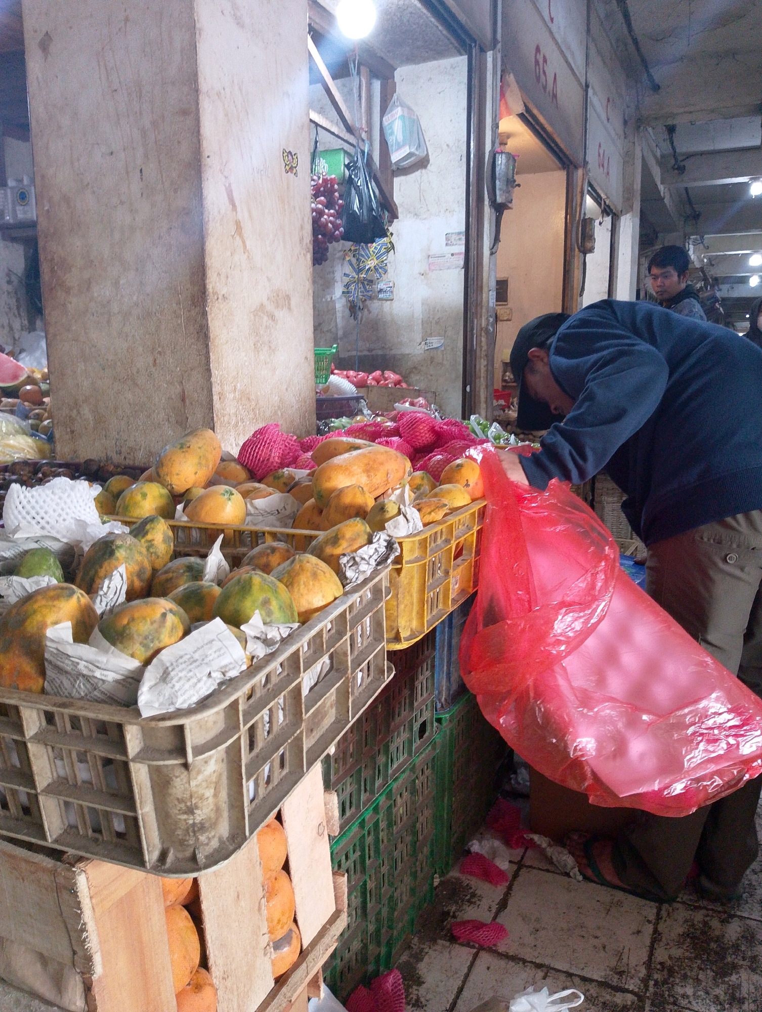 This is a photograph taken in Lembang, Indonesia, depicting a street market scene, likely a fruit stall. In the foreground, a person wearing a dark blue jacket, a black cap, and brown pants is bending over, interacting with a large, translucent red plastic bag that is partially filled with fruit. The bag is positioned in front of a stack of crates filled with papayas. Some papayas are visible in the crates, as well as loose papayas in a wooden crate below. The crates are made of plastic and wood. In the background, more fruit, including what appears to be watermelons and other colorful fruits, are displayed. There are also bunches of red grapes hanging. The environment appears to be an indoor or covered market, with concrete flooring and visible overhead structures. The lighting suggests it is daytime, possibly with artificial lights in addition to natural light. Another individual, a man with dark hair, is partially visible in the background to the right, looking towards the front. The overall impression is of a busy, possibly slightly cluttered, market environment.