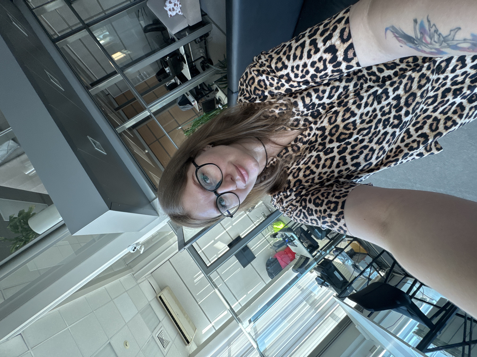 The image is a selfie of a woman wearing black-rimmed glasses and a leopard print blouse. She is positioned in what appears to be an office environment with extensive glass partitions and a multi-story structure visible through the glass. The presence of a plant on a high ledge and various office furnishings suggests an indoor workspace. The location is identified as Perm, Russia, which is based on the presence of specific architectural style and urban landscaping that are typical of Perm.