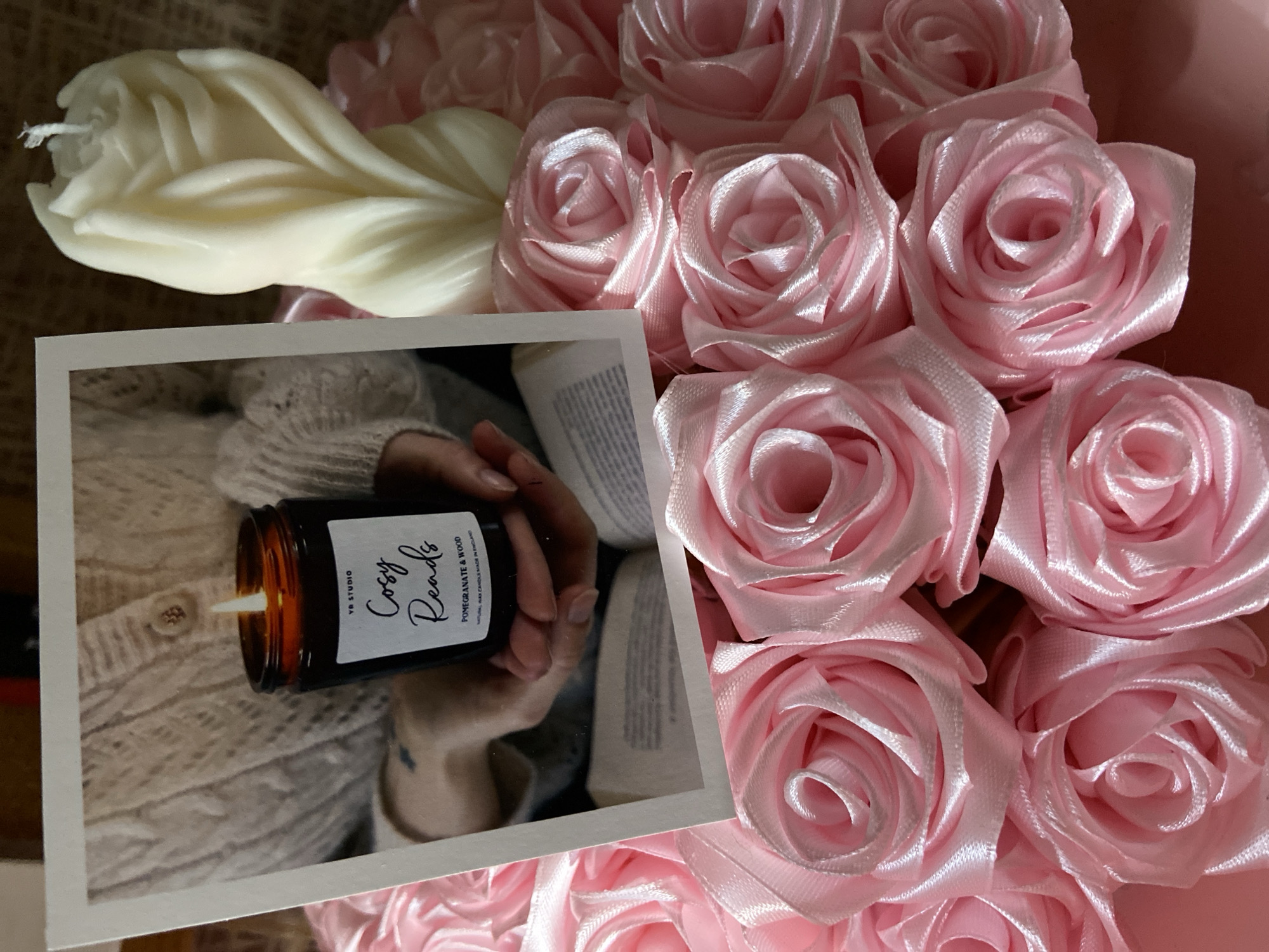 The image shows a close-up of a decorative arrangement featuring pink satin roses and two candles. One candle, in a dark glass jar with a label reading "Cosy Reads," is lit and positioned behind a postcard-like image of a person holding the same candle. The postcard image also shows a book. To the right of the jar candle is a tall, cream-colored candle with a twisted, sculpted design, resembling a flower or flame. The background is a textured brown surface. The arrangement is not located in the city of Япрыково, Russia; no geographical information can be inferred from the image.