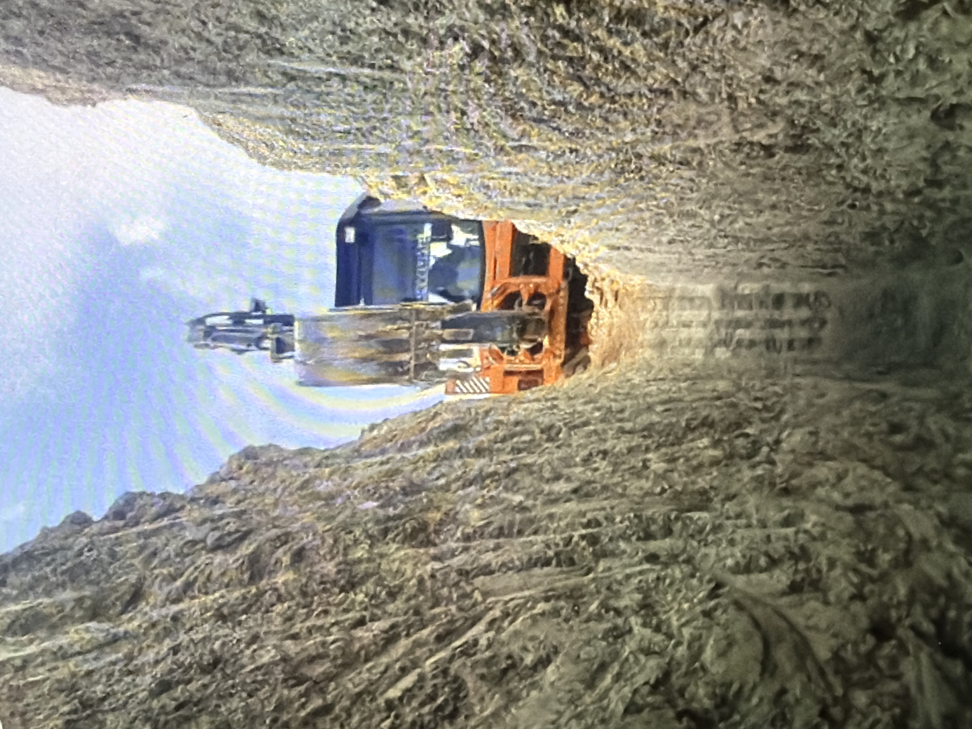In a trench dug into the earth, an orange and black excavator sits with its bucket extended. The trench walls are rough and textured, made of packed dirt and rock. The sky above is blue with scattered clouds. The excavator's cabin is visible, with a dark silhouette of a person inside. The overall scene suggests active construction or excavation work.