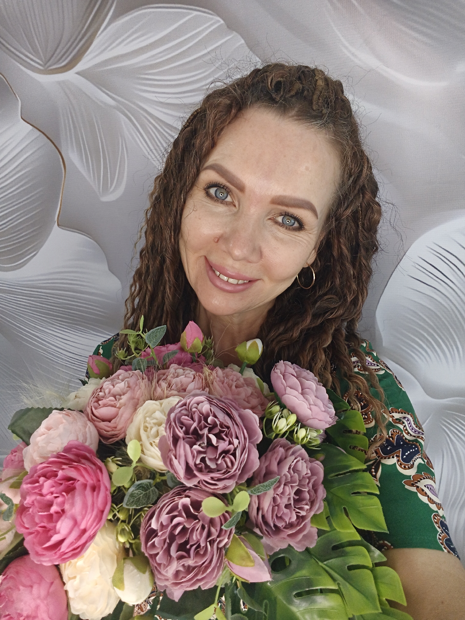 A woman with curly brown hair and blue eyes holds a bouquet of pink and purple roses and green leaves. She wears gold hoop earrings and a green patterned dress. The background is a white wall with a leaf pattern. The location and city are unknown.