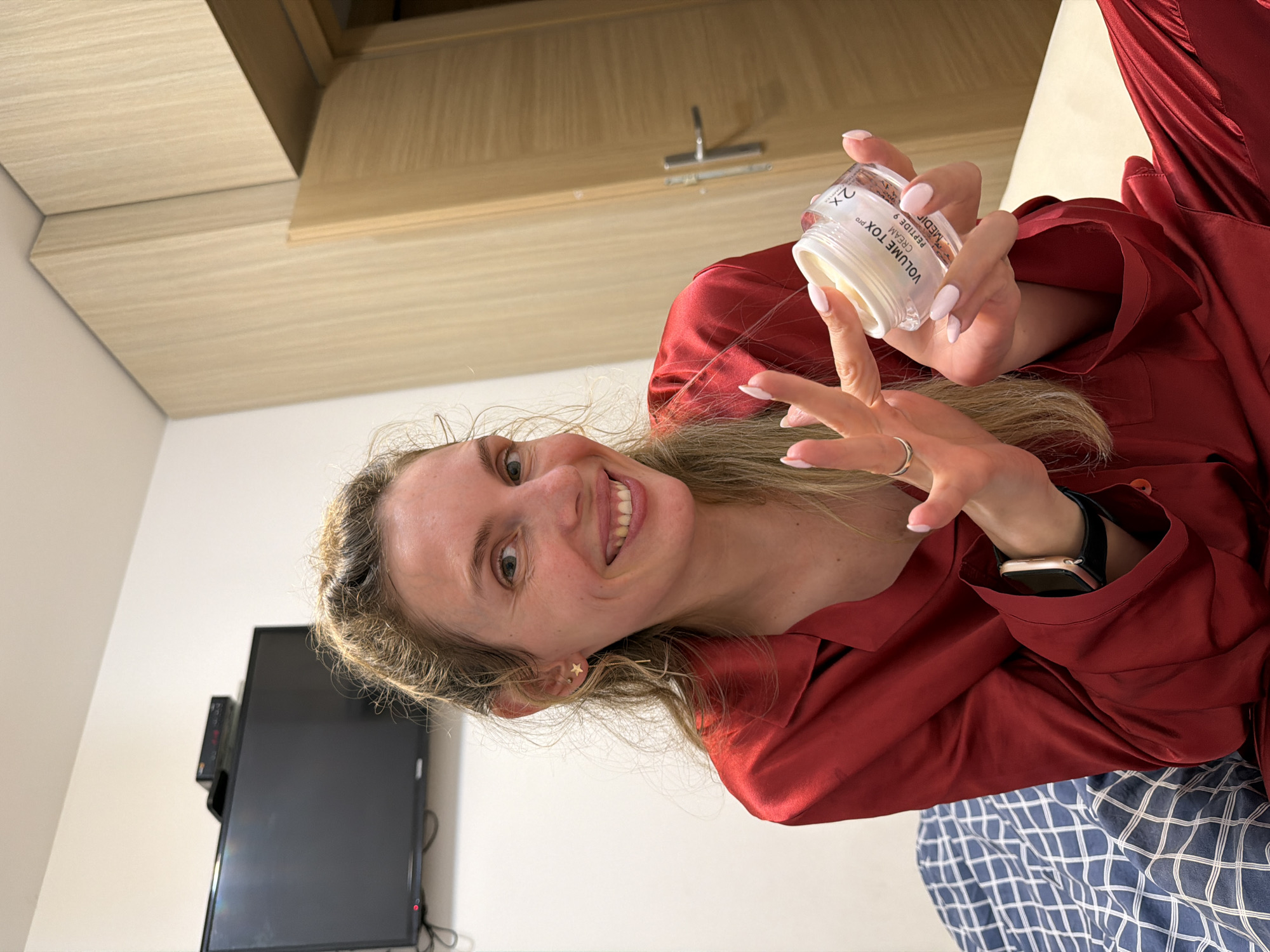 A fair-skinned woman with blonde hair is holding a jar of "VOLUME TOX" cream while sitting on a bed with a blue and white checkered pattern in what appears to be a room in Budva, Montenegro. She is wearing a maroon satin pajama top and an Apple watch. A television is mounted on the wall behind her. The room has light-colored wood paneling on the ceiling and a white wall.