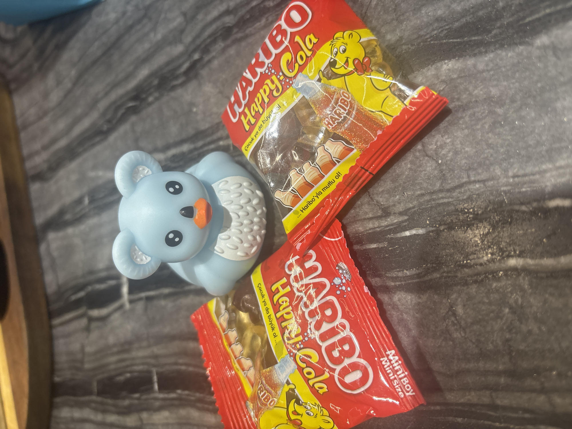 A blue koala-shaped rubber duck is positioned behind two red and yellow-packaged Haribo Happy Cola gummy candies. The candies are laid out on a dark gray speckled countertop. The packaging features images of cola bottles and cartoon bears. The koala duck has large, dark eyes and an orange beak. The lighting suggests an indoor setting. There is no information in the image to determine its location in Dubai, United Arab Emirates.