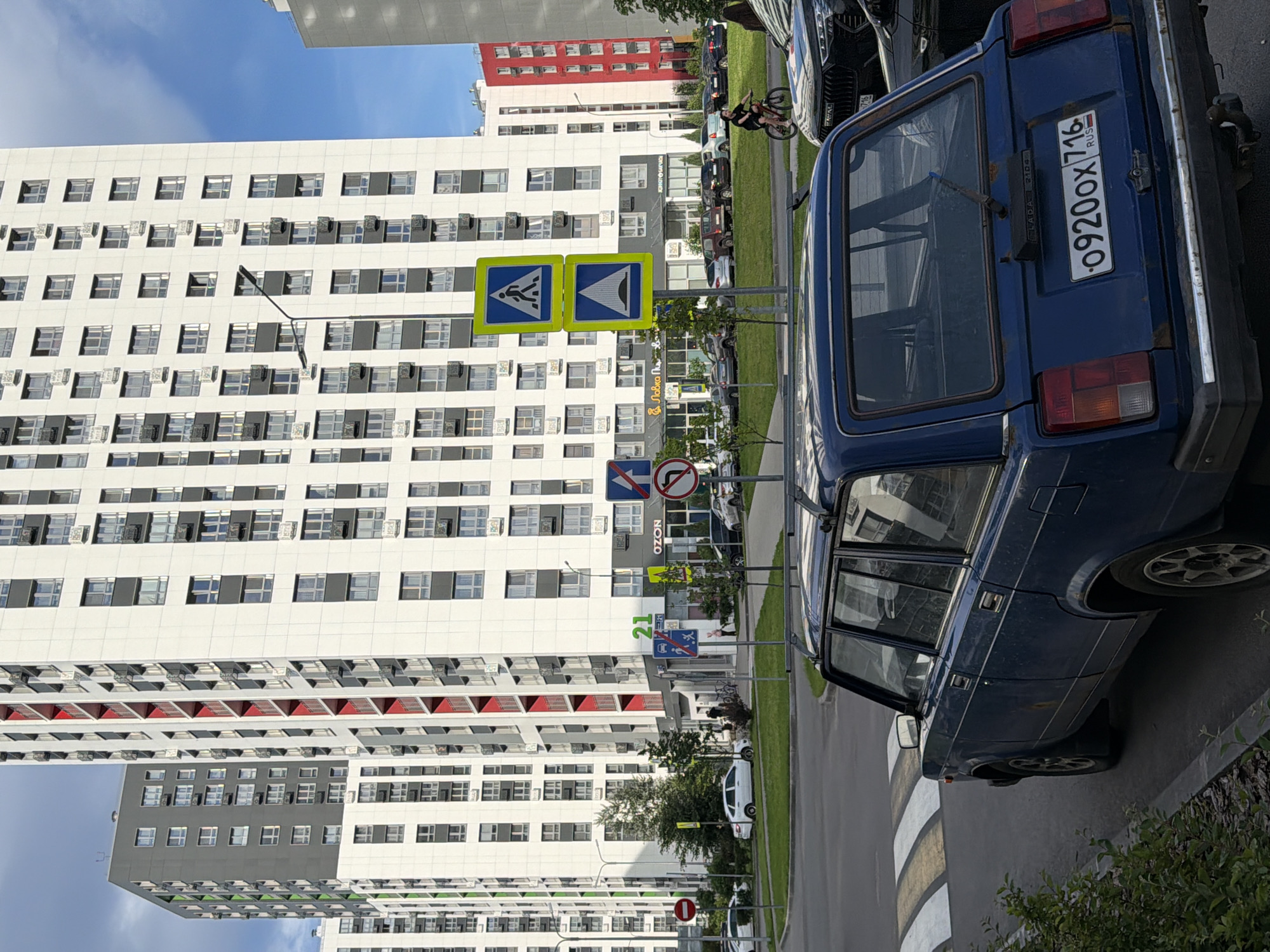 The image depicts a street scene in Kazan, Russia, under a bright blue sky with scattered clouds, suggesting daytime. The primary subjects are two modern, multi-story apartment buildings with white facades and numerous windows, one of which has red accents on its lower levels. A dark blue, older model car, likely a Lada, is parked on the right side of the road, taking up a significant portion of the frame. The car has a visible Russian license plate: "o 9200x 716 RUS".
In the foreground, a crosswalk with distinctive white and yellow stripes is visible. Several traffic signs are present along the roadside. Notably, there are two yellow square signs with blue triangular symbols, one depicting a pedestrian crossing and another showing a ramp or incline, indicating a pedestrian crossing and a potential elevation change or ramp. Other traffic signs, including a red circle with a diagonal line and a symbol of a vehicle, and a blue rectangular sign with a white pedestrian figure and an arrow, are also visible further down the street.
The scene appears to be a residential area. The apartment buildings are the dominant architectural feature, suggesting a developed urban environment. The presence of the car, road, and crosswalk indicates a functioning street. There are no people clearly depicted engaging in specific activities, though a person on a bicycle is visible in the background, near the car. The overall atmosphere is one of a typical urban day. The time of day is clearly indicated by the bright sunlight and blue sky.