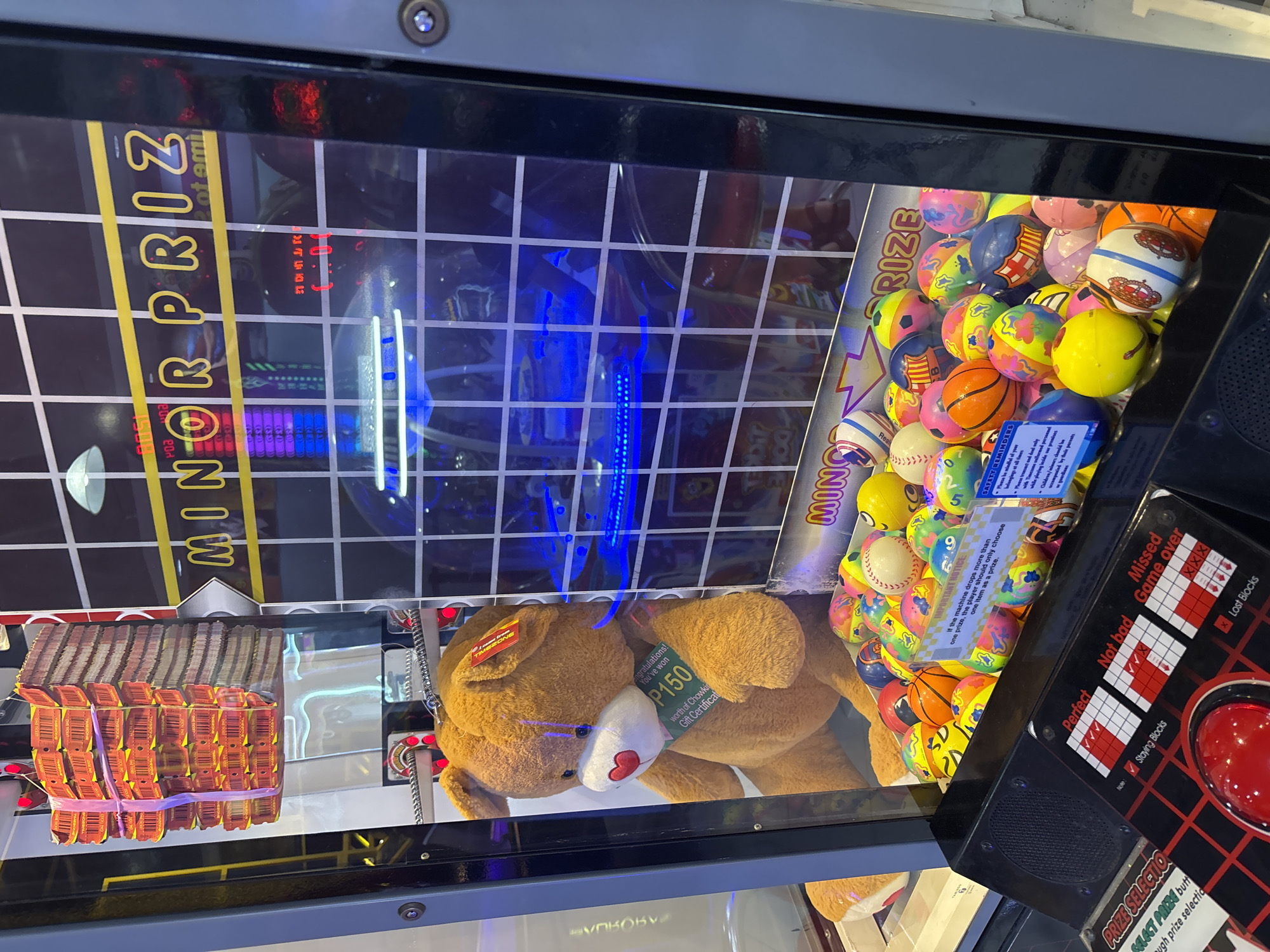 The image shows a claw machine filled with various colorful balls and a large teddy bear. A stack of orange tickets, tied with a pink ribbon, is hanging from the top of the machine. The teddy bear has a white heart-shaped nose and is wearing a green tag that reads "Congratulations! You've won P150 worth of Chowking Gift Certificate". The machine has a grid-like pattern on the back with blue lights illuminating the interior. Text on the machine reads "MINOR PRIZE". A control panel at the bottom displays options for "Perfect", "Not bad", and "Missed" with corresponding point values and an image of blocks. Safety reminders are also visible on the machine. This claw machine is located at a game arcade in Gaisano Mall, Davao City, Philippines.
