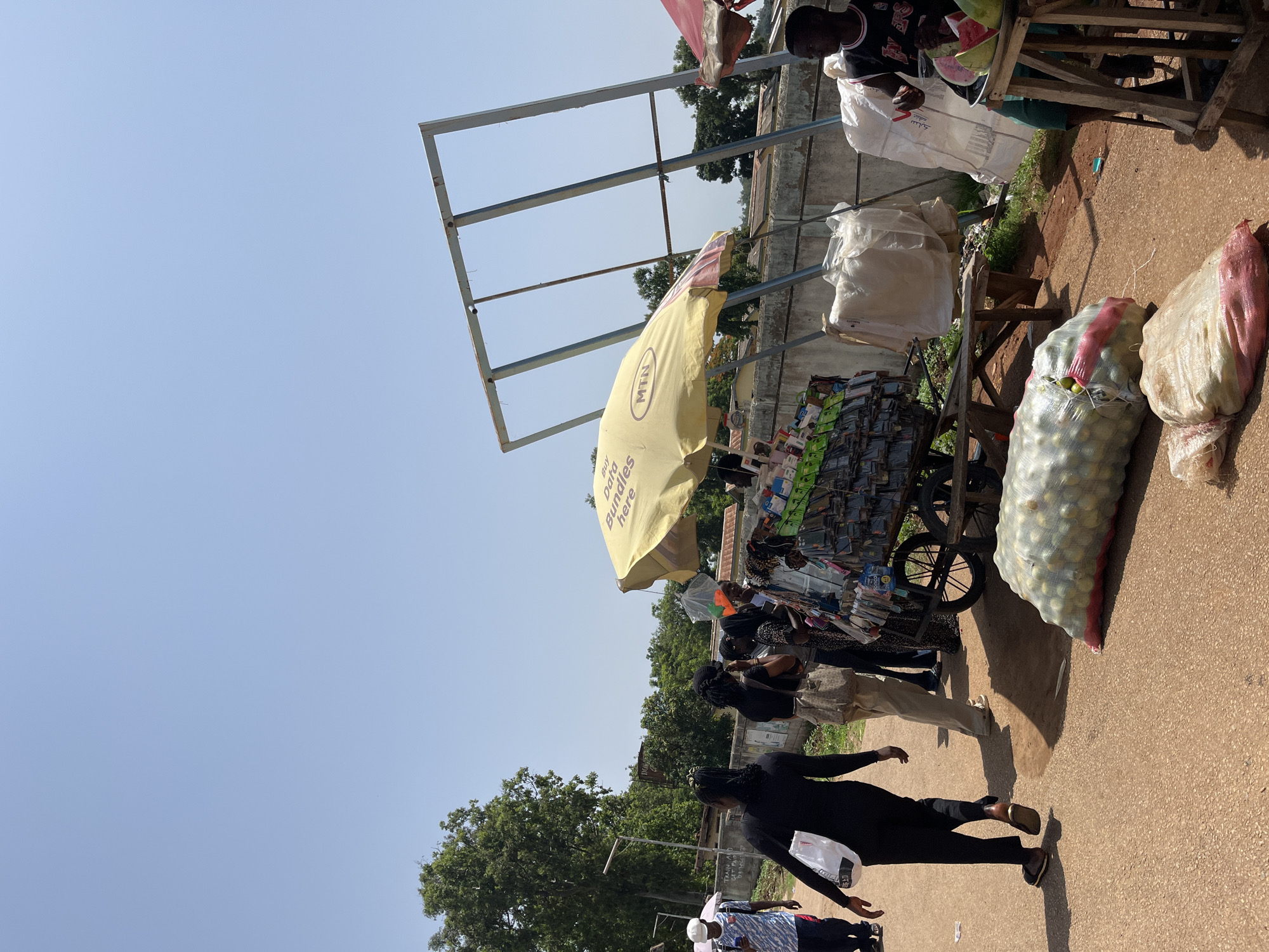 This is a street scene in Rafin Par, Nigeria, with a clear blue sky. On the right side of the image, a vendor is selling fruit, including watermelon and what appear to be small green fruits, possibly limes or passion fruits. He is seated on a wooden stool. Next to him is a stall with several large, white sacks, possibly containing produce. In front of the stall, a cart is laden with bags of produce, likely onions, based on their shape and color. The cart also displays various items for sale under a yellow umbrella, including what appear to be phone accessories.
Several people are walking past the stalls. On the left side of the image, a person wearing a black top and black pants walks away from the camera, carrying a white shopping bag. Behind them, another person in lighter-colored pants walks in the same direction. Further back, a group of people are gathered around the stall with the yellow umbrella, possibly interacting with the vendor or browsing the merchandise. A man in a black tank top with red lettering is standing on the right side, near the fruit stall. In the background, there is a concrete wall and some trees. A large, rusted metal frame stands partially erected against the sky.