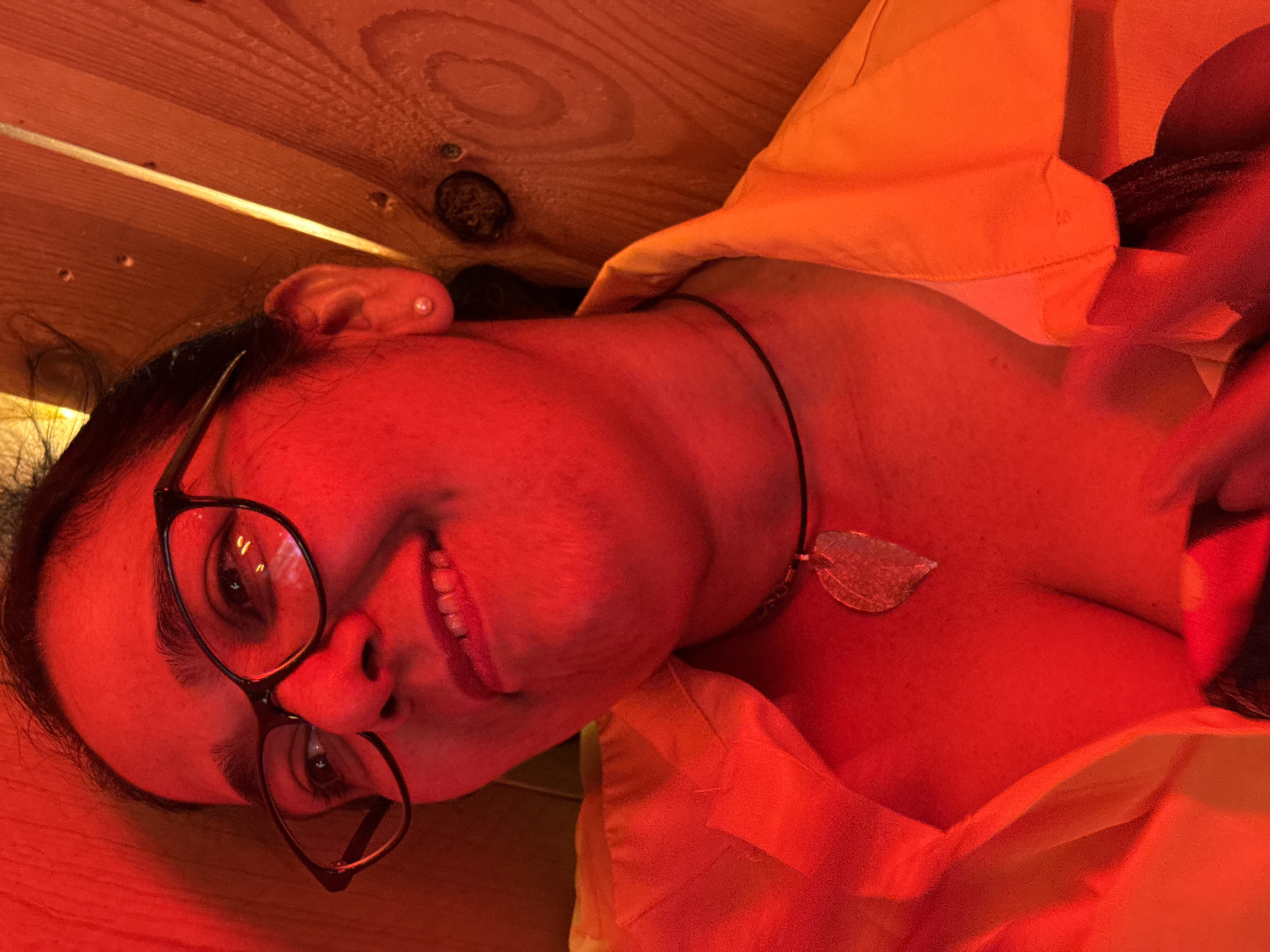 The image shows a close-up, low-angle shot of a person wearing glasses and an orange shirt. The person has dark hair and is smiling, with their teeth showing. They are wearing a necklace with a leaf-shaped pendant and a small earring in their visible ear. The background appears to be made of wooden planks, with warm lighting that gives the scene a red hue. There is no visible indication of the location in Barcelona, Spain, or any specific details about the environment beyond the wooden planks and lighting. The image captures a candid moment, with the person looking up and slightly to the side.